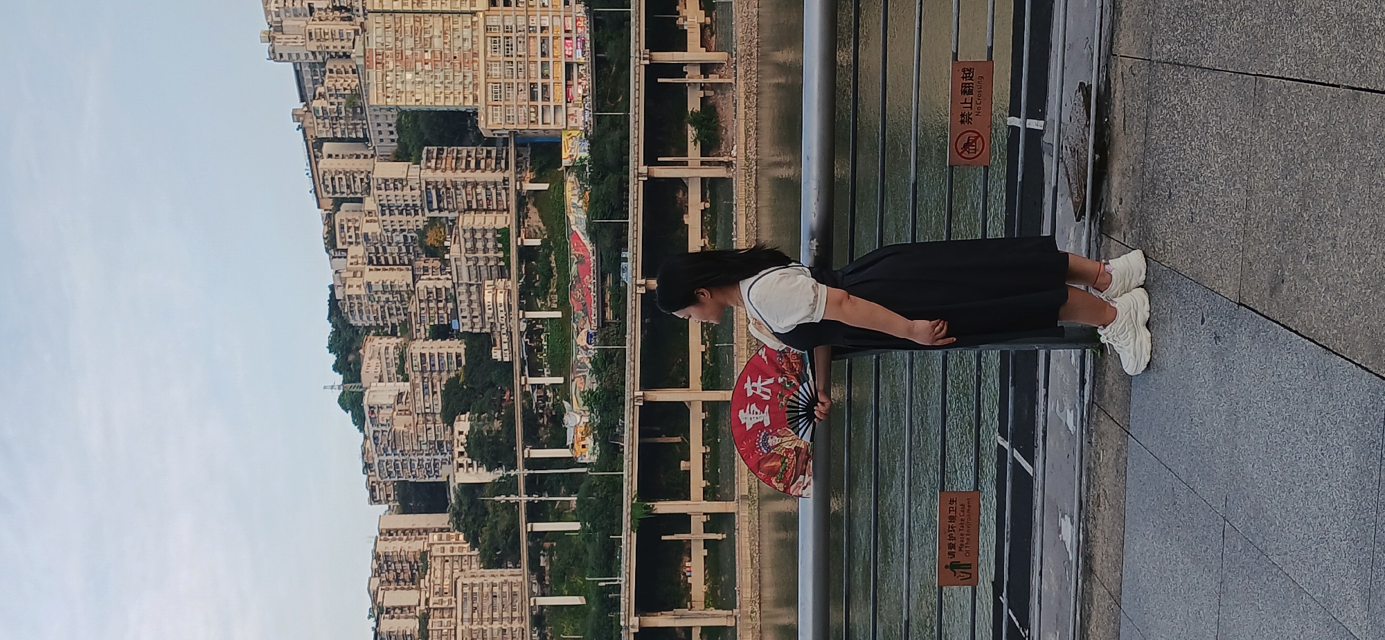 An outdoor, rotated full-shot captures a person standing next to a railing overlooking a river and a densely built cityscape, likely in Jiangbei District, Chongqing, China. The scene is set in what appears to be late afternoon or early evening under a clear sky.
The main subject is a person, appearing to be female, dressed in a white short-sleeved top with a black pinafore-style dress, and white chunky sneakers. She has long, dark hair and is looking down and slightly to her left, towards the river. She holds a large, red, circular fan in her left hand, adorned with traditional Chinese artwork depicting a figure and the Chinese characters "重庆" (Chóngqìng), which refers to the city.
Beyond the railing, a wide river flows, flanked by a complex urban landscape. Numerous multi-story residential and commercial buildings are stacked on a hillside, characteristic of Chongqing's mountainous topography. Various bridge or overpass structures are visible across the river and leading up to the urban areas. Greenery is interspersed among the buildings, particularly on the lower slopes.
Two signs are attached to the railing. One sign, with a brown background, displays Chinese characters "请爱护环境卫生" (Please Take Care Of The Environment) and the English translation "Please Take Care Of The Environment," accompanied by an icon of a person discarding trash into a bin. Another sign, with a red border, reads "禁止翻越" (No Crossing) and "No Crossing" in English, with a prohibition icon showing a person attempting to climb over a barrier. The person's relaxed posture suggests she is enjoying the view or posing for a photograph along a waterfront promenade.