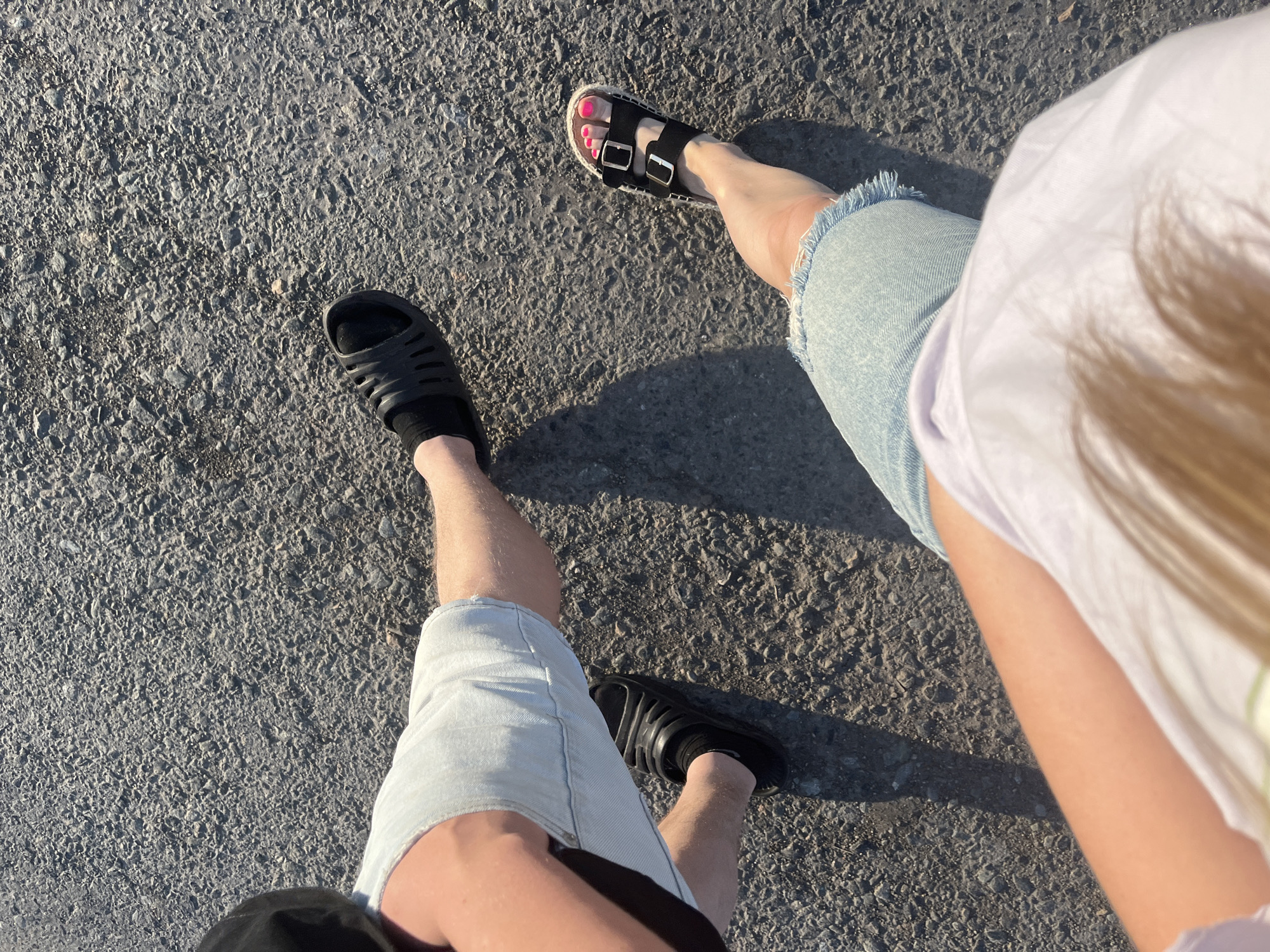 The image shows a top-down view of two people's legs walking on an asphalt surface. The person on the left is wearing light blue denim shorts and black slide sandals with black socks. The person on the right is wearing similar light blue denim shorts and black platform sandals with buckles, revealing bright pink toenails. Both individuals appear to be in motion, casting shadows on the textured gray asphalt. The lighting suggests it is daytime, likely outdoors. The environment is a paved surface, consistent with a road or walkway. There is no visible indication of the location being specifically within the city of Mal'tsevo, Russia, or any other identifiable urban area.