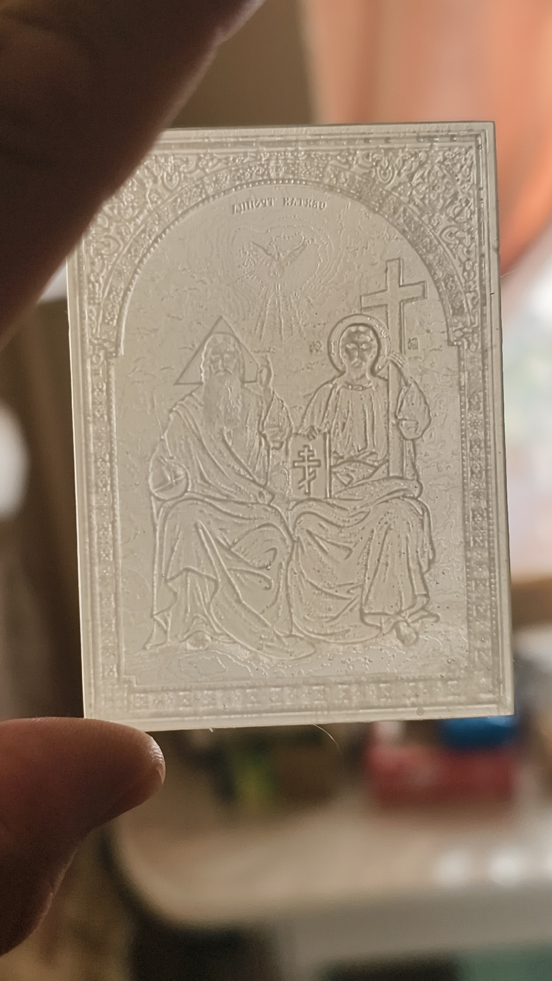 A close-up, eye-level, horizontal shot shows a person holding up a translucent, rectangular devotional item. The item appears to be made of plastic or glass and features an embossed image of the Holy Trinity. The central figures are seated and facing forward. On the left is an older figure with a long beard, possibly representing God the Father, with a triangle shape above his head. To the right is a younger figure with a halo, likely Jesus Christ, holding a book with a cross on its cover. Above them, a dove with outstretched wings symbolizes the Holy Spirit. A cross is depicted to the right of the seated figures. Decorative borders frame the scene. The item is held by fingers visible in the foreground. The background is blurred and appears to be an indoor setting with hints of furniture and other objects.