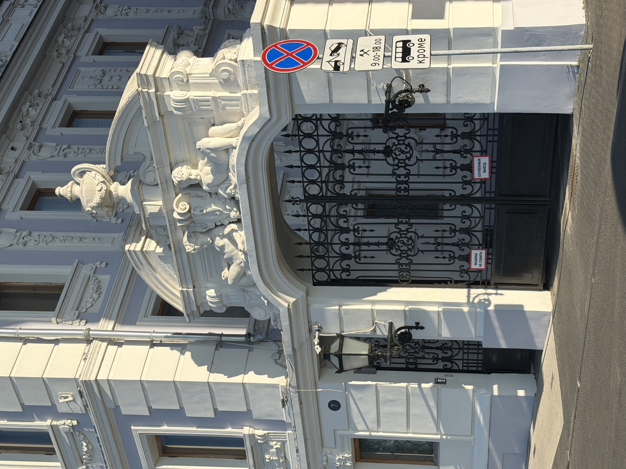 The image shows the facade of a building with ornate architectural details, including sculptures above an arched gate. The building is painted in light blue and white, with decorative moldings and reliefs. A black wrought-iron gate is the central feature of the entrance. A street lamp with a lantern is mounted to the left of the gate. Several traffic signs are visible on the right side of the gate. One sign prohibits stopping, and below it are signs indicating towing, working hours from 9:00 to 18:00, and a bus symbol with the word "кроме" (except). Red signs are attached to the gate, one reading "МАШИНЫ НЕ СТАВИТЬ!" (Do not park cars!) and another reading "СЛУЖЕБНЫЙ ВЫЕЗД" (Service exit). The image appears to be taken during daylight, with shadows cast by the building and its architectural elements. The location is identified as the Kustarev House at 7 Likhachev Street, Nizhny Novgorod, Russia.