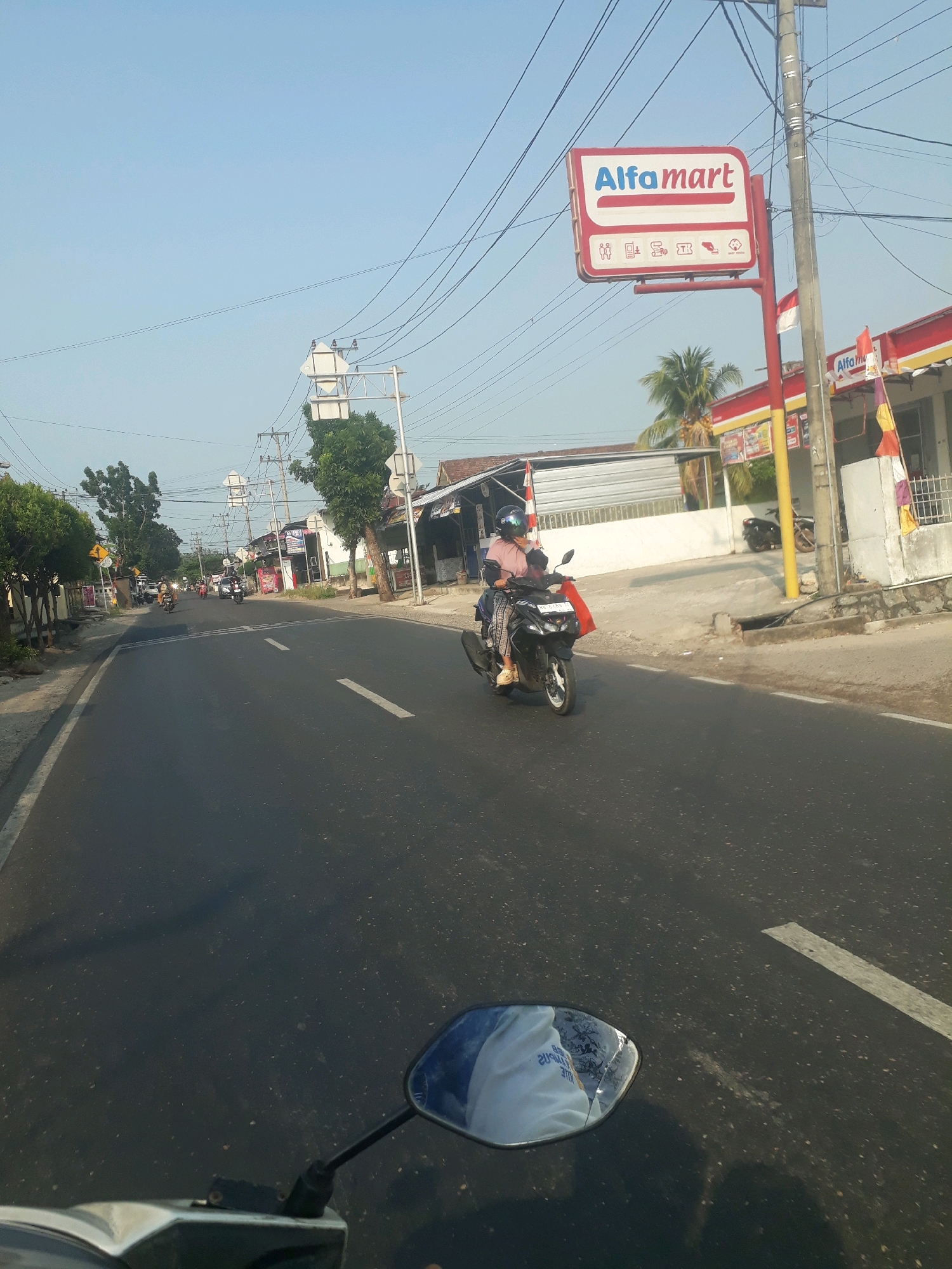 The image depicts a street scene in Pangkalpinang, Indonesia, under a clear blue sky. A prominent "Alfamart" sign, red and white, is visible on the right side of the road, mounted on a pole. The sign features the Alfamart logo and several icons representing services. A streetlamp and power lines crisscross overhead.
In the foreground, the reflection of a person wearing a white shirt with blue text is visible in the rearview mirror of a motorcycle, suggesting the photo was taken from a motorcycle's perspective. The road itself is asphalt with white lane markings.
Further down the road, several motorcycles are traveling in the same direction. One motorcycle in the middle ground carries a rider wearing a helmet and a pink shirt, with a red bag attached to the back. To the right of the road, there are buildings, including an Alfamart store with a red and yellow awning, and a palm tree. The left side of the road features trees and other structures. The overall impression is of a typical street scene in a Southeast Asian city.