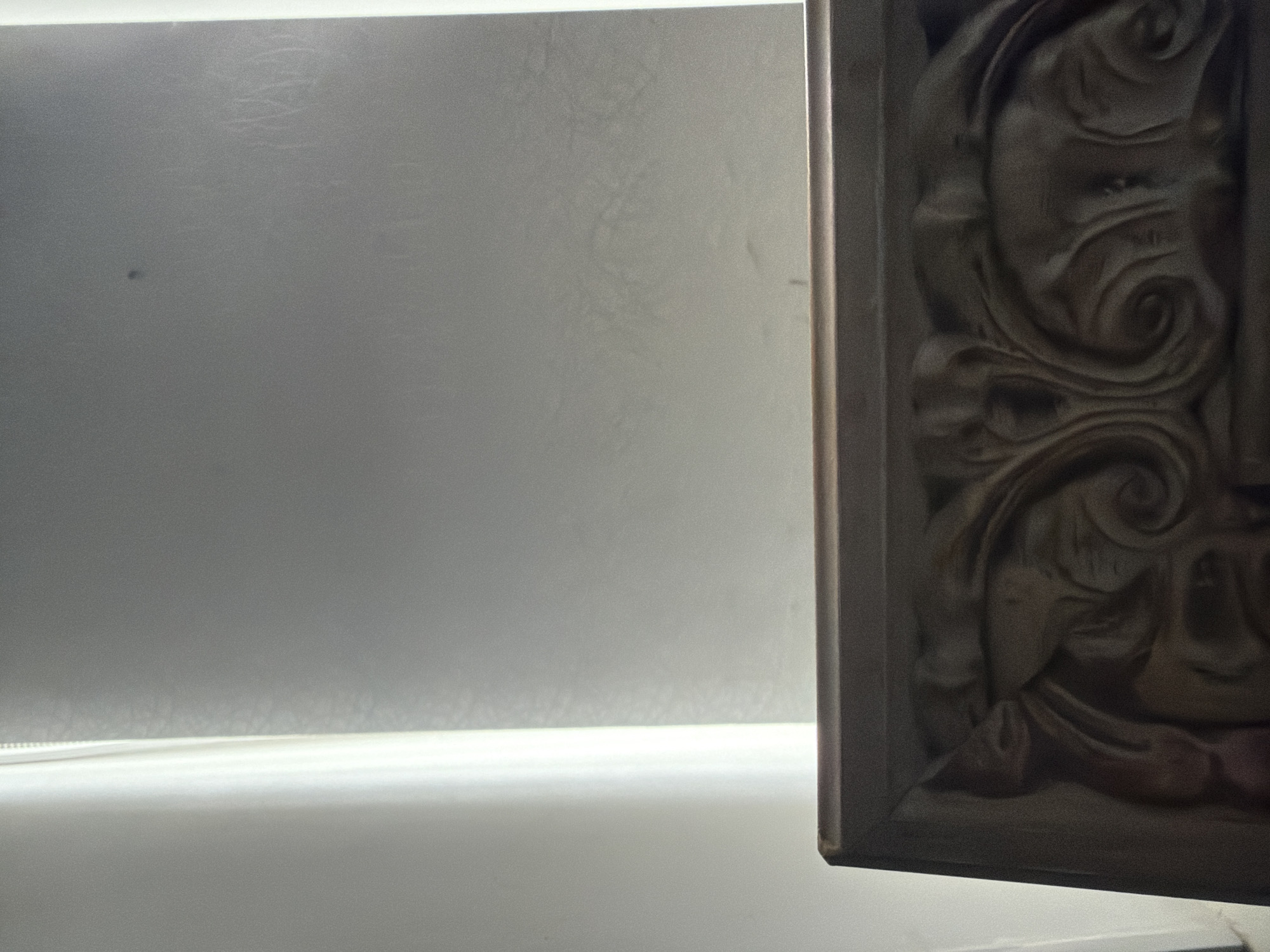 A close-up shot focuses on the ornate carvings of what appears to be a wooden frame or furniture. The carving features intricate swirling patterns and what could be stylized faces or floral motifs. The wood is dark and appears to have a matte finish, with some areas catching the light. In the background, a bright light source shines from the left, possibly a window or lamp, casting a soft glow and creating a gradient of light on a textured surface. The overall impression is one of detail and craftsmanship.