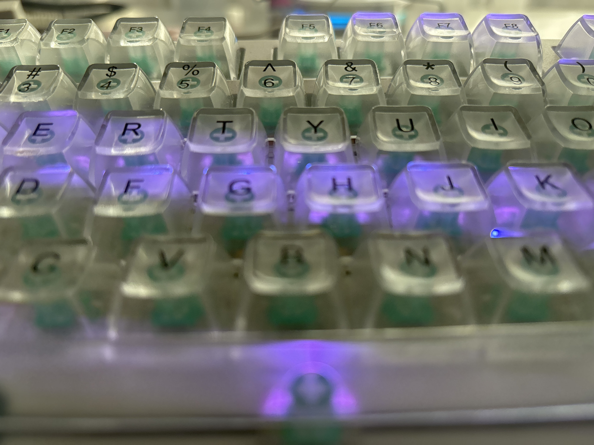 The image is a close-up, eye-level shot of a white mechanical keyboard with translucent keycaps that glow with purple and blue backlighting. The keyboard is the sole subject of the image, and it is positioned horizontally. The keycaps have letters and symbols printed on them. In the top row, functional keys labeled F1 through F8 are visible. Below that, number keys with associated symbols are present, such as #3, $4, %5, ^6, &7, *8, and (9. The rows below show letter keys including E, R, T, Y, U, I, O, P, D, F, G, H, J, K, L, and various symbols. The bottom row shows keys like C, V, B, N, and M. The lighting from the keyboard creates a vibrant, illuminated effect on the surrounding surface, which appears to be a desk or table. The background is out of focus, suggesting a shallow depth of field. The overall scene is well-lit, with the primary illumination coming from the keyboard itself.