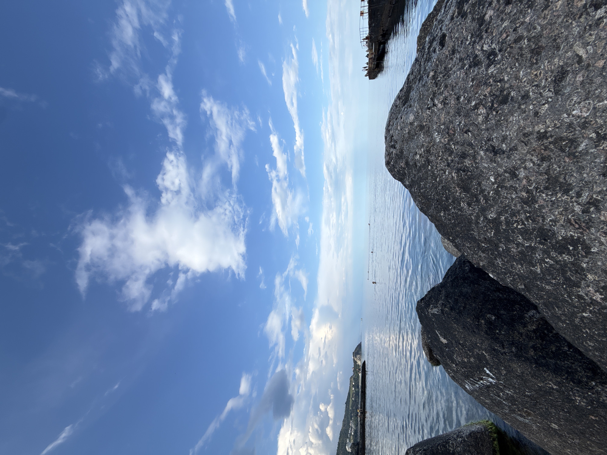 The image depicts a tranquil scene at the sea. In the foreground, large, dark gray rocks dominate the composition, partially submerged in the clear, rippling water. The water stretches out to the horizon, meeting a vast blue sky dotted with scattered white clouds. In the middle ground, a distant shoreline with buildings and greenery is visible on the left, while a weathered concrete pier extends into the sea on the right, with a few silhouetted figures standing on it. Several other figures are scattered in the water, seemingly swimming or enjoying the sea. The overall impression is one of a peaceful day by the water.