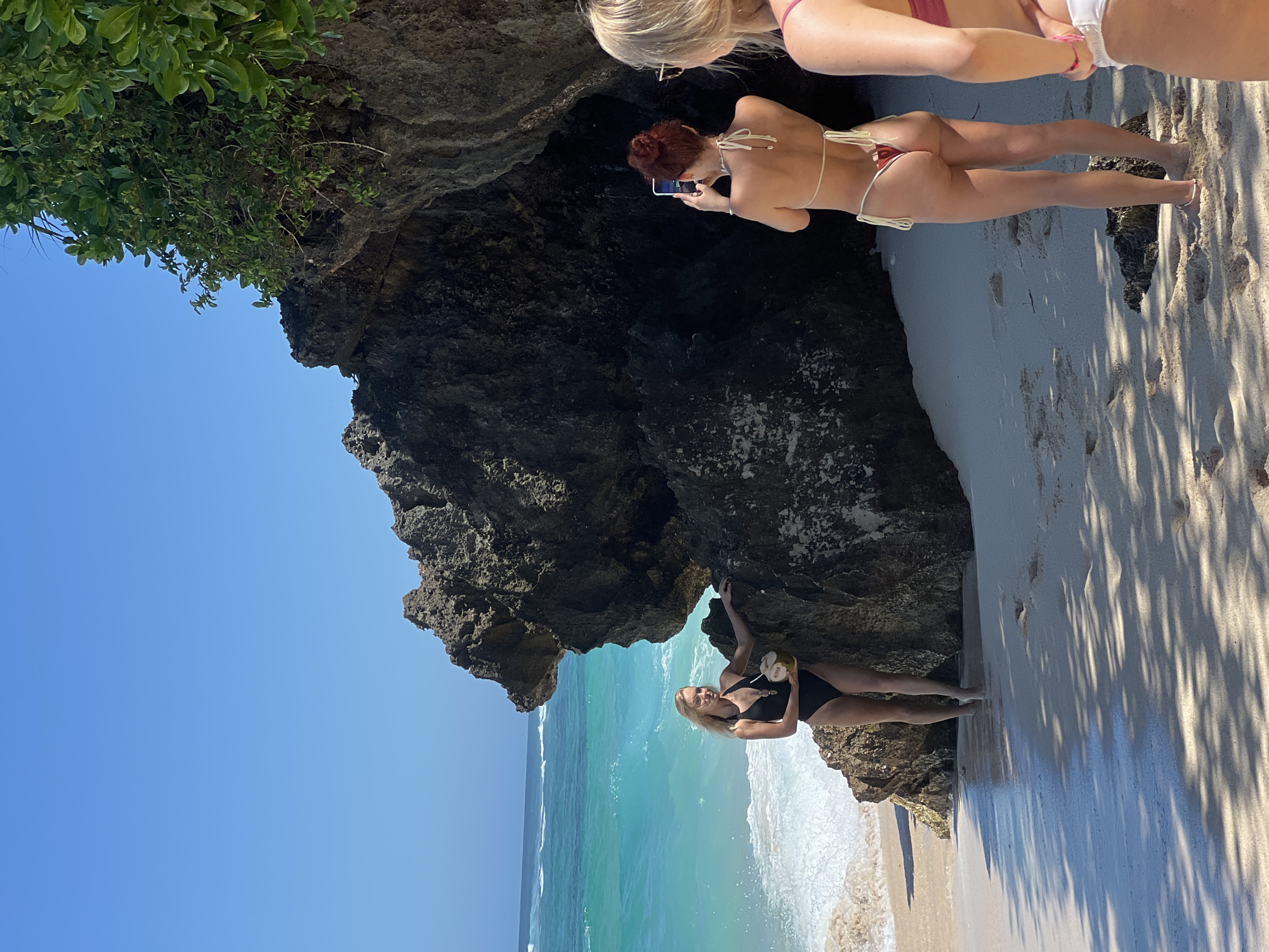 The image depicts a coastal scene in Kuta Selatan, Indonesia. Dominating the view is a large, dark rock formation that forms a small cave or overhang on a beach. Two women are positioned near the formation; one wearing a black swimsuit is holding a coconut. Another woman in a bikini stands in the cave, taking a picture with a cell phone. The sand is light-colored and stretches along the shoreline, with tree shadows stretching over the surface of the sand. The ocean is turquoise in color.
