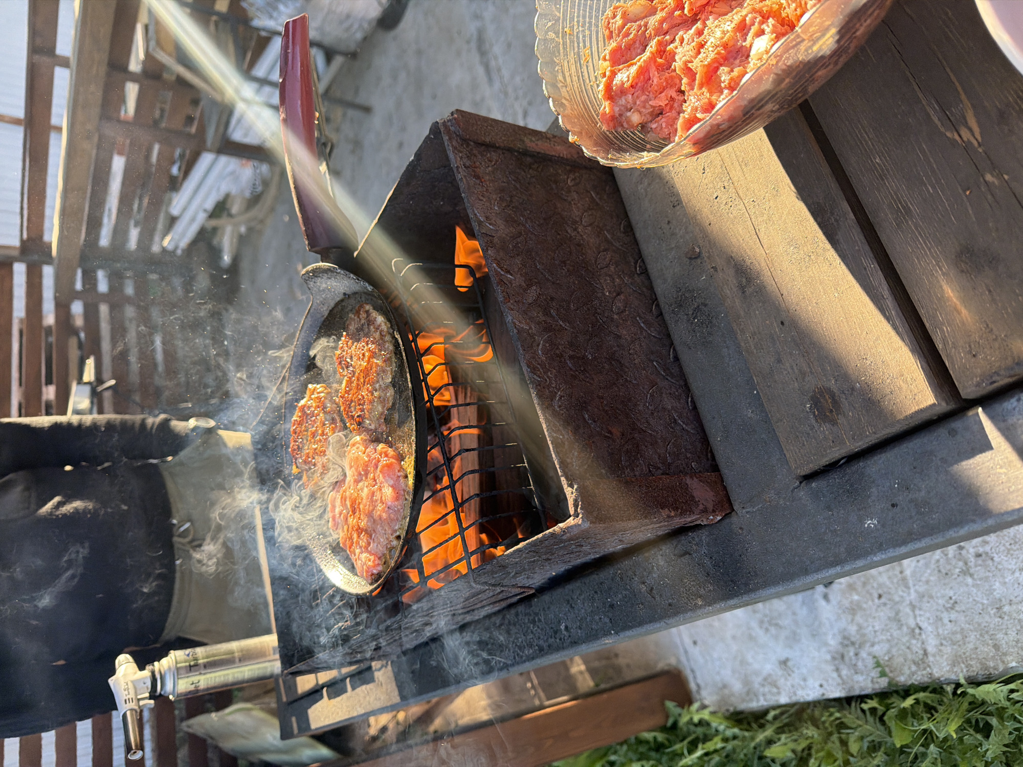 A bowl of raw meat sits on a wooden table next to a grill that has meat cooking on a pan above a fire. A hand holding a culinary torch is near the pan. The location appears to be outside in daylight, near some greenery. The available information indicates this may be in Большая Шильна, Russia.