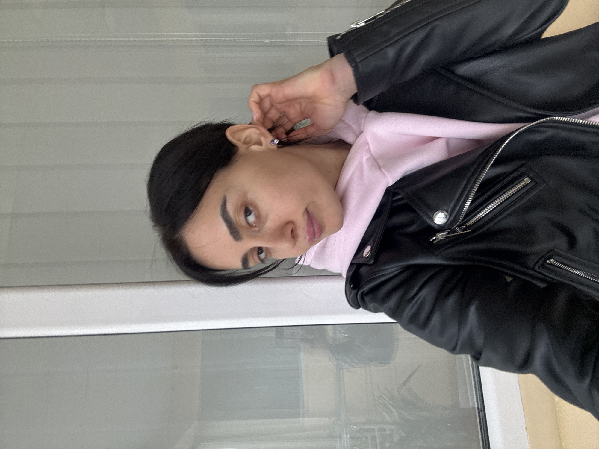 A woman is visible in this portrait-style shot. She has dark hair and is wearing a black leather jacket over a pink hoodie. She is touching her ear. The background appears to be a window with some out-of-focus greenery visible through it. There is no explicit evidence in the image to confirm the location. The location is stated to be Tbilisi, Georgia.