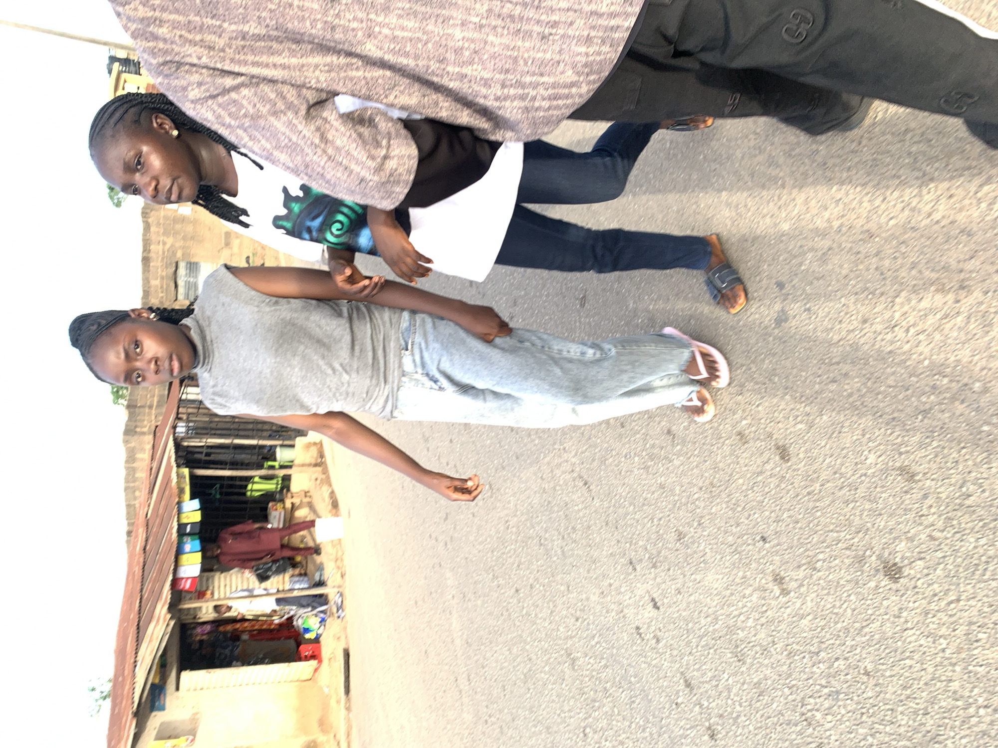 The image shows three individuals on a paved road, with a building featuring corrugated metal roofing visible in the background, typical of the architecture in Jos, Nigeria. One individual is wearing a grey turtleneck shirt and light blue jeans. Another is partially visible, wearing a white t-shirt with a design, a black shirt, and dark blue jeans. The third individual is mostly cut off from the top of the image.
