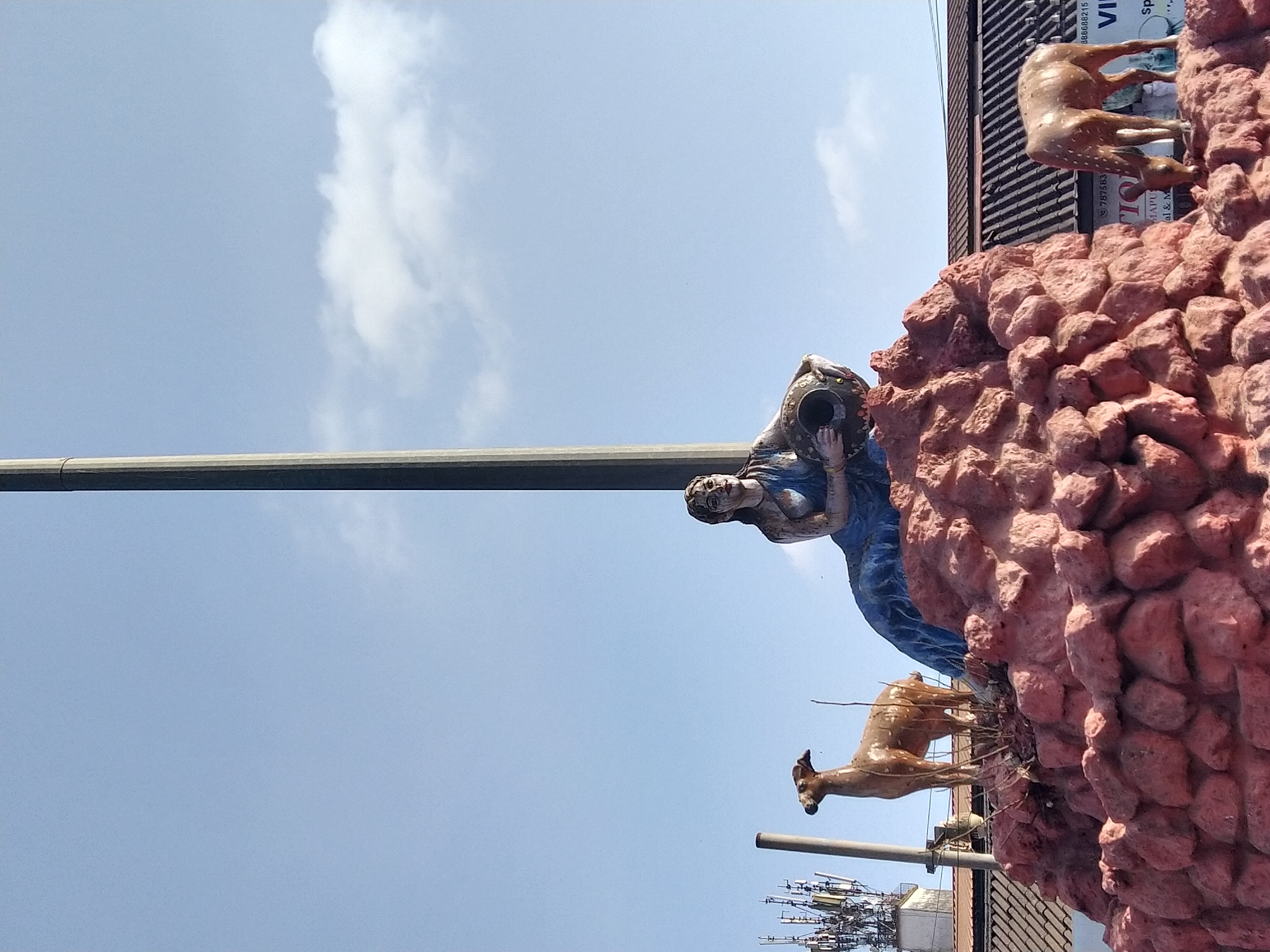 The image shows an outdoor statue in Mapusa, India. The statue features a woman in blue clothing holding a vessel, deer, and a red, rock-like structure, all set against a partly cloudy sky. Utility poles and building tops are visible below.