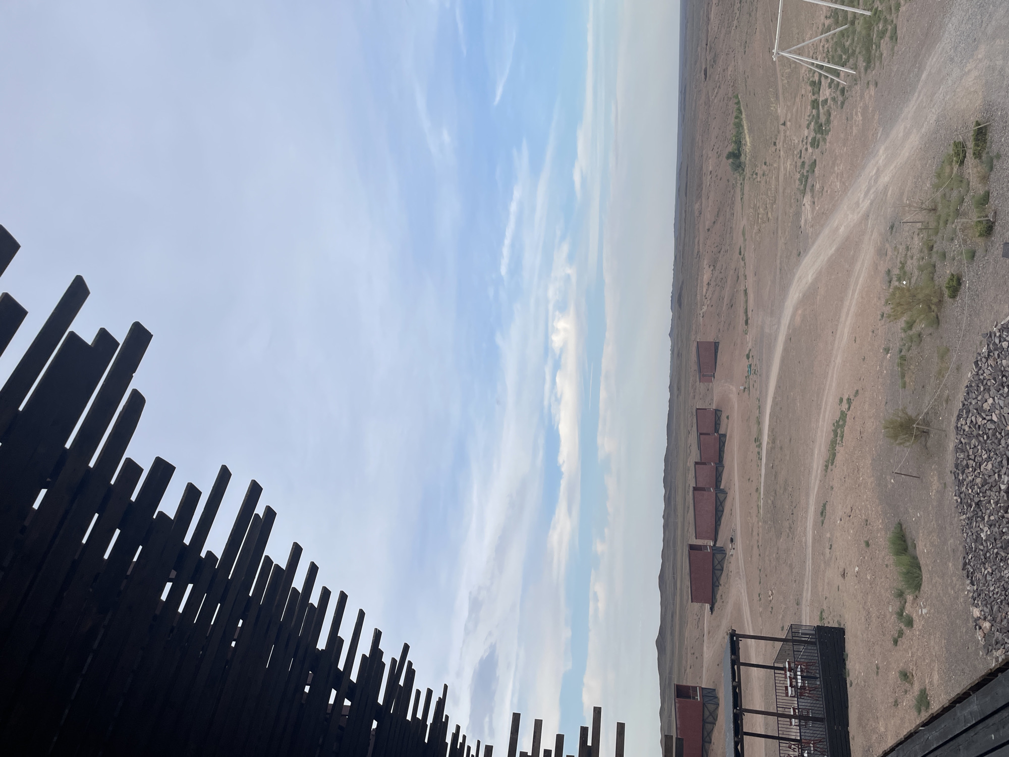 A perspective shot from a dark wooden structure with layered slats shows a sprawling, arid landscape under a pale blue sky with scattered white clouds. In the mid-ground, a series of identical, reddish-brown rectangular structures with angular supports are arranged in a line across the dusty terrain. A dirt road winds through the landscape, passing by scattered low-lying green vegetation. To the left, a dark, covered outdoor seating area with tables and chairs is visible, and a pile of grey stones is in the foreground. To the right, a white swing set structure stands against the distant, rolling hills. The scene appears to be a resort or unique lodging in a remote, natural environment, consistent with locations like the town of Tamerlik, Kazakhstan.