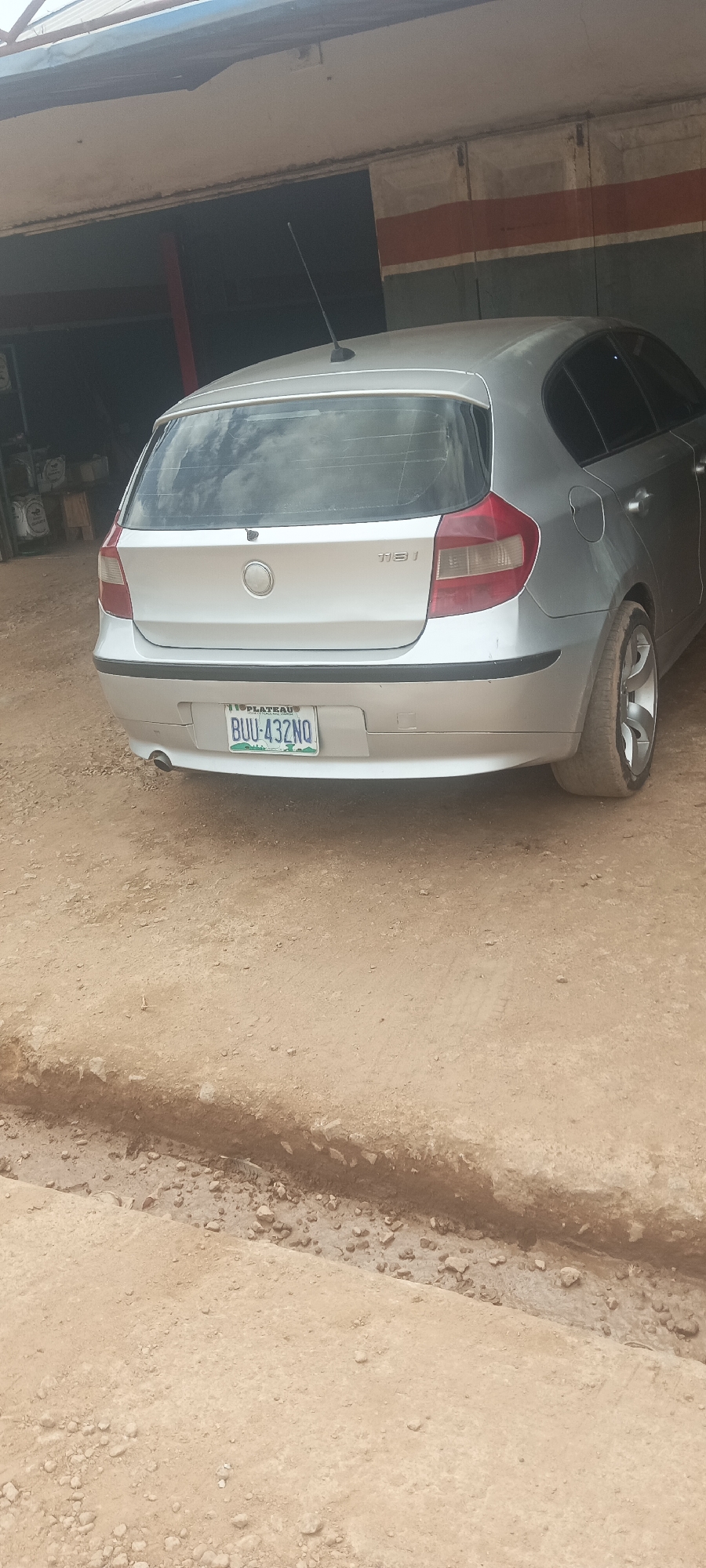 A silver BMW 1 Series hatchback is parked on a dirt surface. The rear of the car is facing the camera, and the license plate is visible. The car is positioned in front of an open-fronted building with a corrugated roof and a banded wall. The ground is uneven and appears to be a construction site or an unpaved area. The overall scene suggests an outdoor setting in a possibly undeveloped or construction area. The license plate reads "BUU-432NQ". The car has a telescopic antenna on its roof.