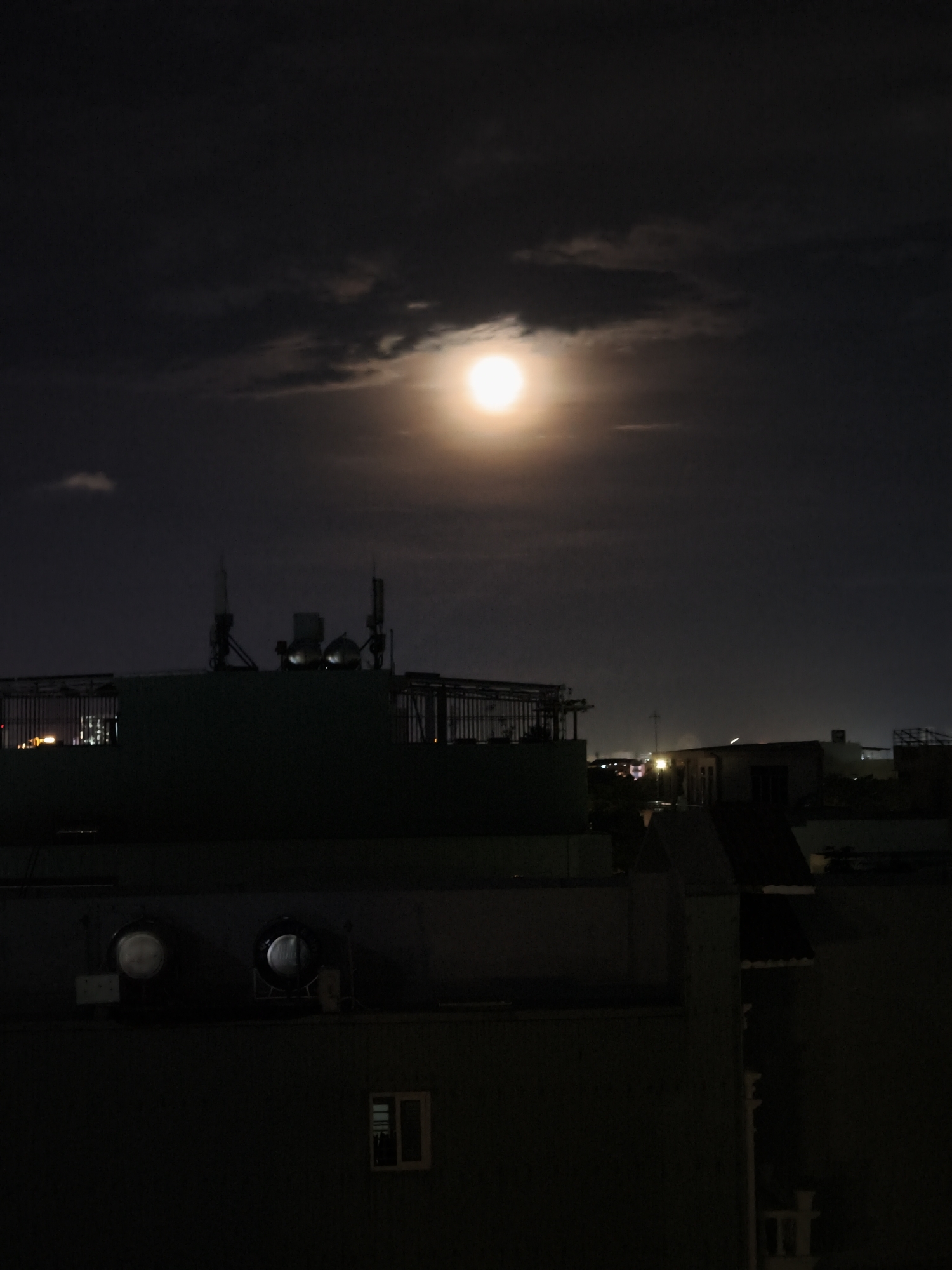 This is a vertical, low-angle shot of a night cityscape, likely in Da Nang, Vietnam. A bright, full moon dominates the upper portion of the frame, its light diffused by dark, wispy clouds. The moon is positioned in the center-right of the sky. Below the moon, silhouetted against the dark sky, are the rooftops of several buildings. Prominent structures include what appear to be communication towers with antennas on one building, and two large, metallic spherical objects on another.
The foreground is filled with the dark, imposing shapes of buildings, some with visible windows and rooftop structures. In the mid-ground, a cluster of buildings shows illuminated windows and what could be streetlights, suggesting an urban environment. The overall mood is dark and somewhat dramatic due to the strong contrast between the bright moon and the deep shadows of the city. The time of day is clearly nighttime, and the weather appears to be partly cloudy. There is no visible text in the image.