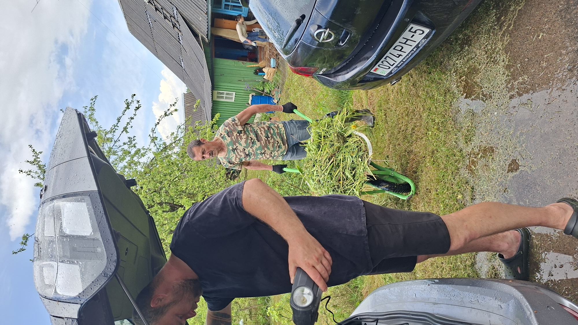 In Сваткаўскі сельскі Савет, Belarus, two men are engaged in outdoor activities. One man, wearing a camouflage shirt and black gloves, is pushing a green wheelbarrow filled with cut grass. The other man, dressed in a black t-shirt and shorts, is holding a black flashlight and appears to be inspecting the open hood of a dark-colored van. A black Opel car with a Belarusian license plate (0274 PH-5) is parked nearby. The setting is a grassy yard with a wooden house visible in the background. The ground appears wet, possibly after a recent rain.
