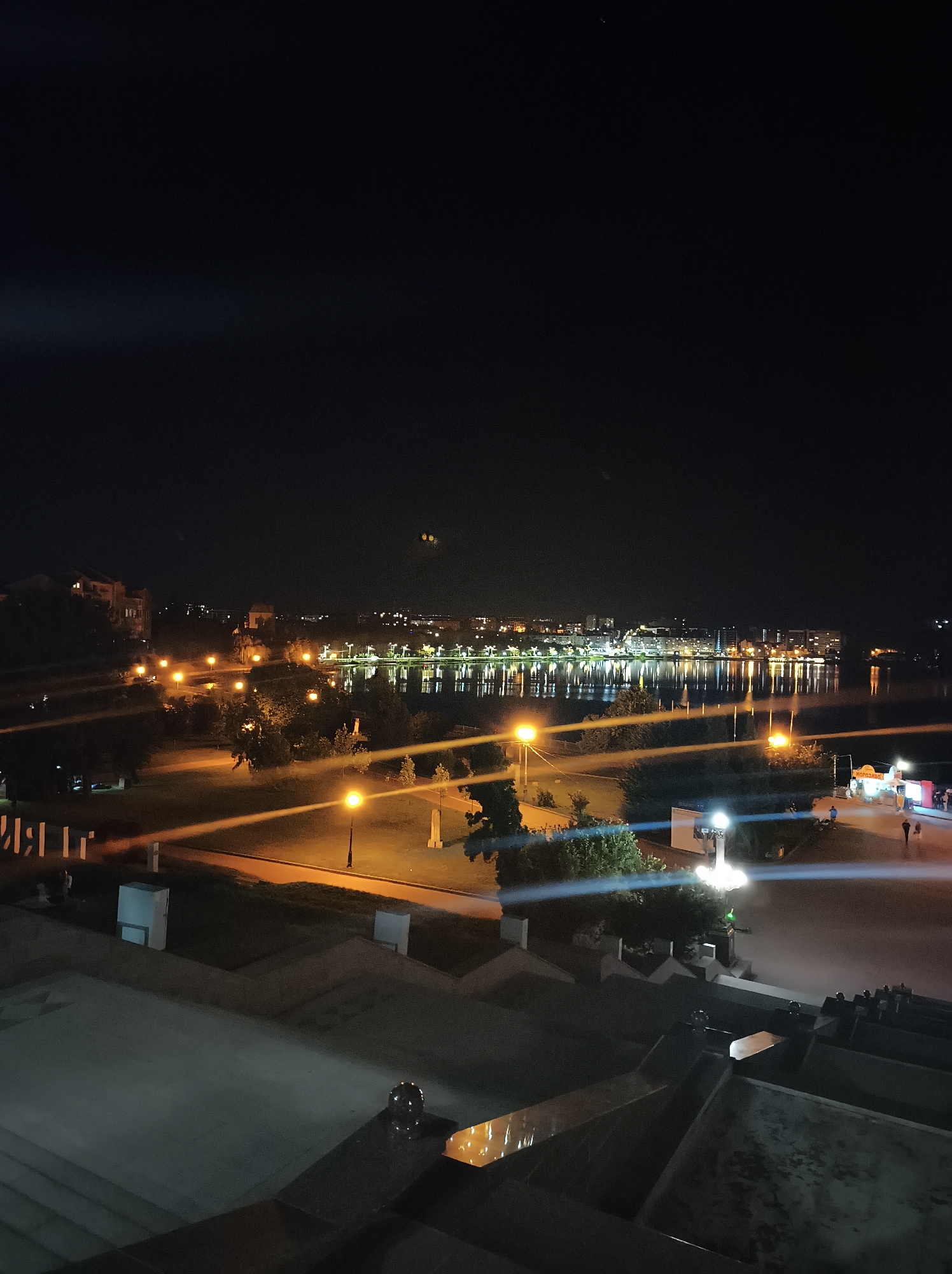This is a nighttime aerial view of the Ternopil Lake in Ternopil, Ukraine. The lake is illuminated by lights from the opposite shore, creating reflections on the water. A park area with walking paths and lampposts is visible on the near side of the lake. There are some buildings and trees in the background. In the foreground, there are large concrete steps and structures. The overall atmosphere is calm and serene.