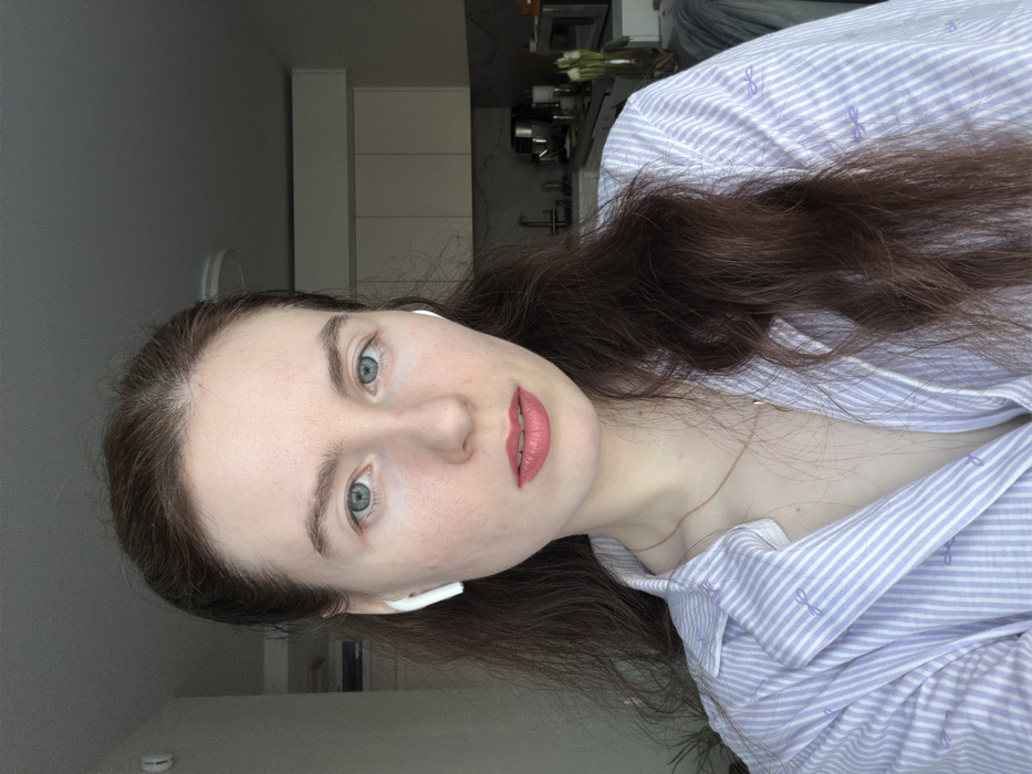 This image captures a chest-up portrait of an adult female in an indoor residential setting, specified as Ковалёво, Russia. The individual, with fair skin, blue-green eyes, and long, dark brown hair styled with a central part, is looking directly at the camera with a neutral expression. She is wearing two white wireless earbuds and has a light pink or coral lipstick applied. Her attire consists of a light purple and white striped collared shirt, partially unbuttoned, revealing a thin rose gold-colored necklace. Small purple bow motifs are visible on her shirt. The background to the viewer's left features a light-colored wall with a partial view of a green potted plant. To the viewer's right, a modern kitchen area is discernible, equipped with light-colored upper cabinets, a dark countertop, a stainless steel stand mixer, a sink, a vase containing white flowers (likely tulips), and a portion of a microwave oven. A circular ceiling light fixture is visible in the upper background. Natural light appears to illuminate the scene from the left.