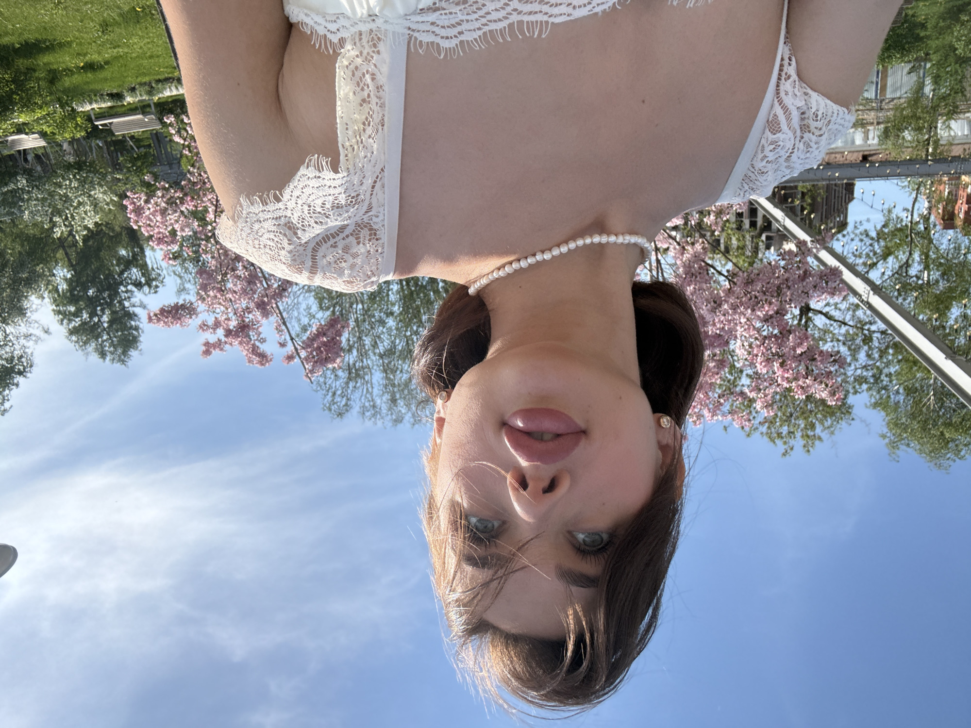 A woman with light skin and dark hair wears a white lace garment and a pearl necklace. The photograph is taken from a low angle, looking up at the woman, who appears to be standing outdoors. Behind her, pink blossoms suggest a flowering tree, and a blue sky is visible. In the background, blurred elements indicate a possible urban park setting, with trees and distant buildings. Based on the visible details, the location cannot be confidently determined to be in Perm, Russia.