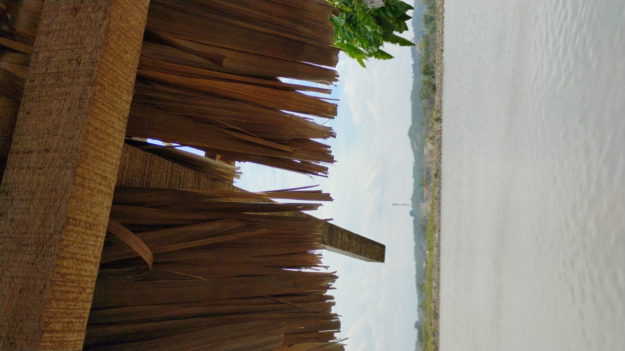 The image is a low-angle shot looking out from under a thatched roof structure towards a body of water. The roof is made of dry palm fronds that are frayed and partially broken, allowing glimpses of the sky. A wooden beam is visible supporting the roof. In the distance, across the water, is a shoreline with a low line of trees and hills. A prominent cell tower is visible on one of the hills. The water appears calm with a slight ripple on its surface. The sky is partly cloudy. The image appears to be taken in Lhokseumawe, Indonesia, suggested by the presence of tropical vegetation and the overall landscape.