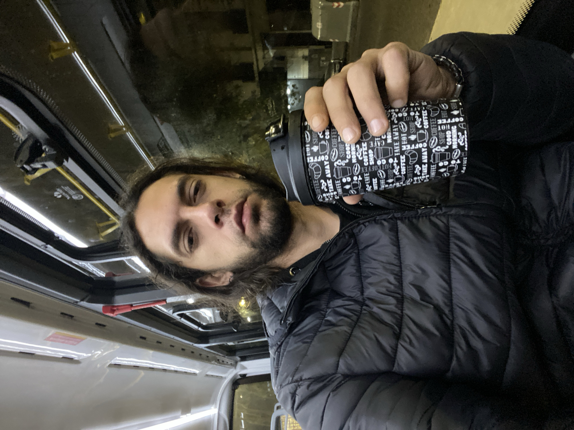 A man with long brown hair and a beard is holding a coffee mug in his right hand. The mug is black with white coffee-themed graphics. He is wearing a black puffer jacket and a bracelet. He is inside a vehicle, possibly a bus, given the overhead emergency exit and interior fittings. Outside, the scene appears to be urban and at night. The location is Buenos Aires, Argentina, although no particular neighborhood is visible. The man appears to be taking a selfie.