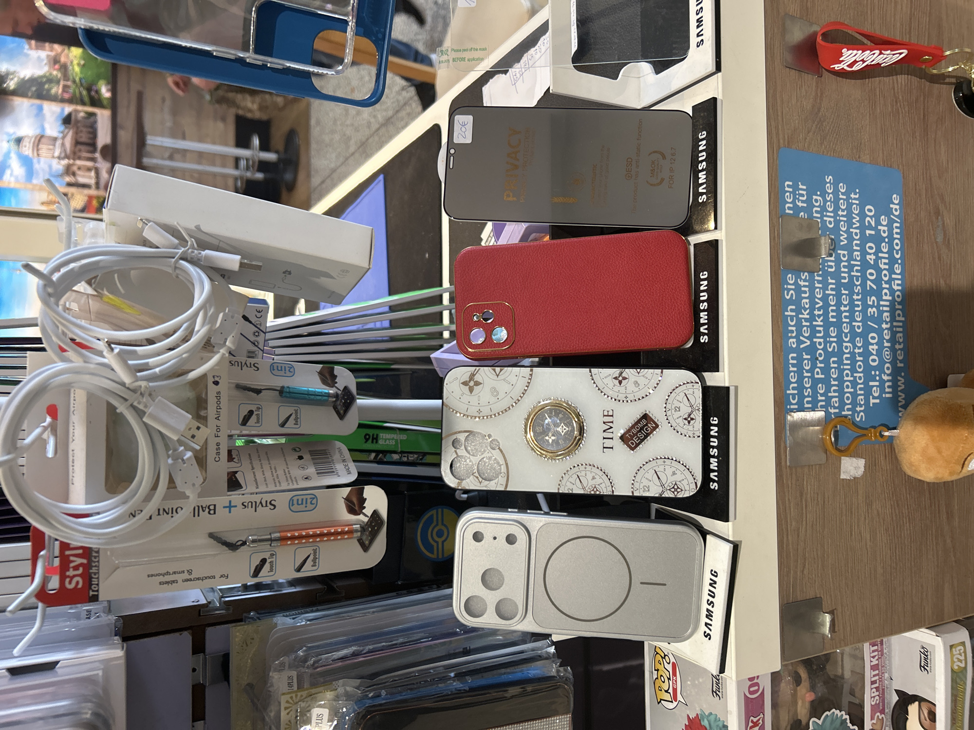The image captures a close-up view of a retail display, likely in a shop or stall, showcasing a variety of mobile phone accessories and other small merchandise. The setting appears to be indoors, under artificial lighting.
In the upper-middle and right sections, several phone screen protectors are visible, including one marked "PRIVACY PROTECTION" and "FOR IP 12 6.7", indicating it's for an iPhone 12. A price tag of "20€" is attached to one. Below the screen protectors are various phone cases: a red leather-textured case, a white case decorated with clock faces and gears with "TIME" and "TYBOMB DESIGN" text, and a silver case with a distinct camera array cutout and a circular magnetic attachment area. "SAMSUNG" branding is visible on the display stands for these items.
To the left, a coiled white charging cable is prominent, alongside packaging for "Case For Airpods 3". Further left, several "2in1 Stylus + Ball Joint" pens are displayed, with features like "Touch Tip" and "Ballpoint", and indications that they are "For touchscreen tablets & smartphones" and "MADE IN CHINA".
Other items include a red "Coca-Cola" keychain, a small brown plush toy/keychain, and packaging for "Funko POP!" figures, specifically mentioning "GPK" and "SPLIT KIT". A blue informational card on the right provides German text, including "retailprofile.com/de" and a telephone number (040 / 35 70 40 120), confirming the retail context and a German-speaking location, aligning with Berlin, Germany. The display appears well-stocked, offering accessories for various popular electronic devices.