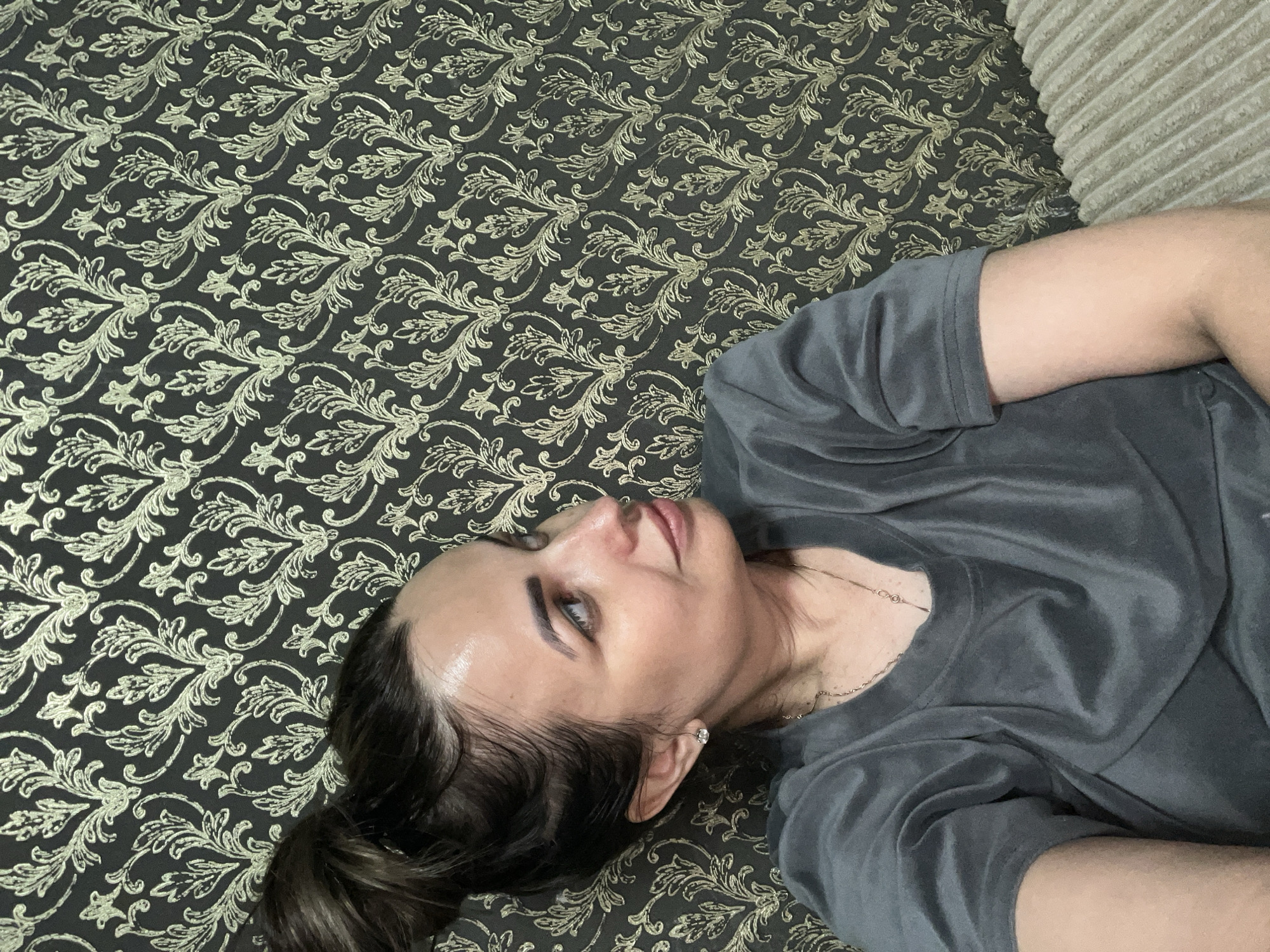 A woman with dark hair pulled back is lying down on a patterned surface. She is wearing a grey t-shirt and a silver necklace. Her head is tilted to the side, and she is looking up and to her left. Her right arm is bent at the elbow, resting near her head, and her left arm is out of frame. The background is a dark fabric with a repeating gold floral pattern. To the right of the patterned fabric, there is a ribbed, beige object that appears to be a cushion or part of a sofa. The lighting suggests an indoor setting, possibly during the daytime. The woman's expression is relaxed and contemplative. There is no visible text in the image.