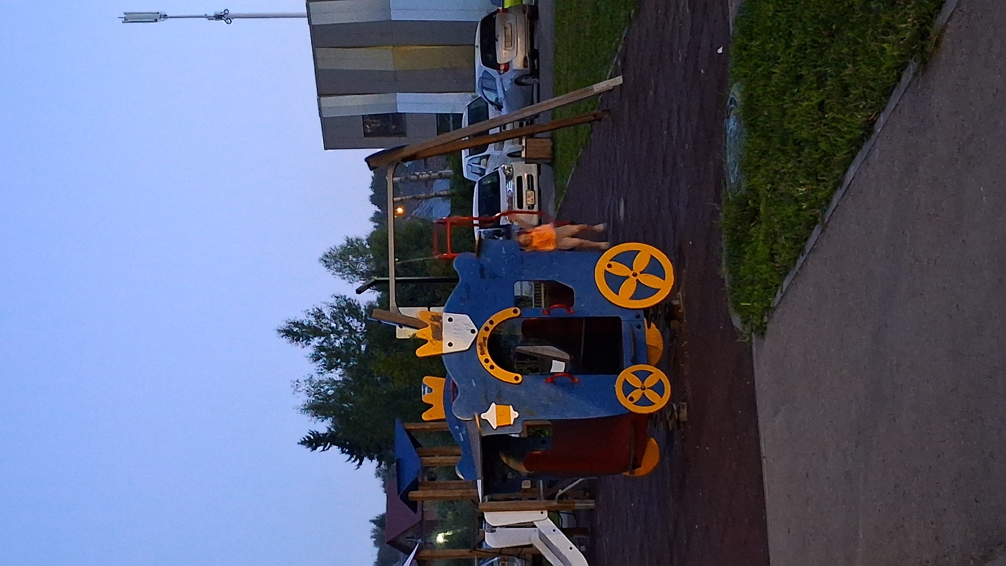 The image depicts a children's playground in the city of Novosibirsk, Russia, likely during dusk. In the foreground, a blue and yellow train-themed play structure is the central focus. A young child, wearing an orange vest, is standing near the structure, with their right arm extended upwards. Behind the train structure, a swing set with a red ladder is visible. The playground is surfaced with dark material, possibly rubber mulch or asphalt. In the background, several parked cars are visible, including a white van and a silver sedan. Beyond the cars, trees and residential buildings are present, with a cell tower rising in the distance. The sky is a pale blue, indicating twilight.