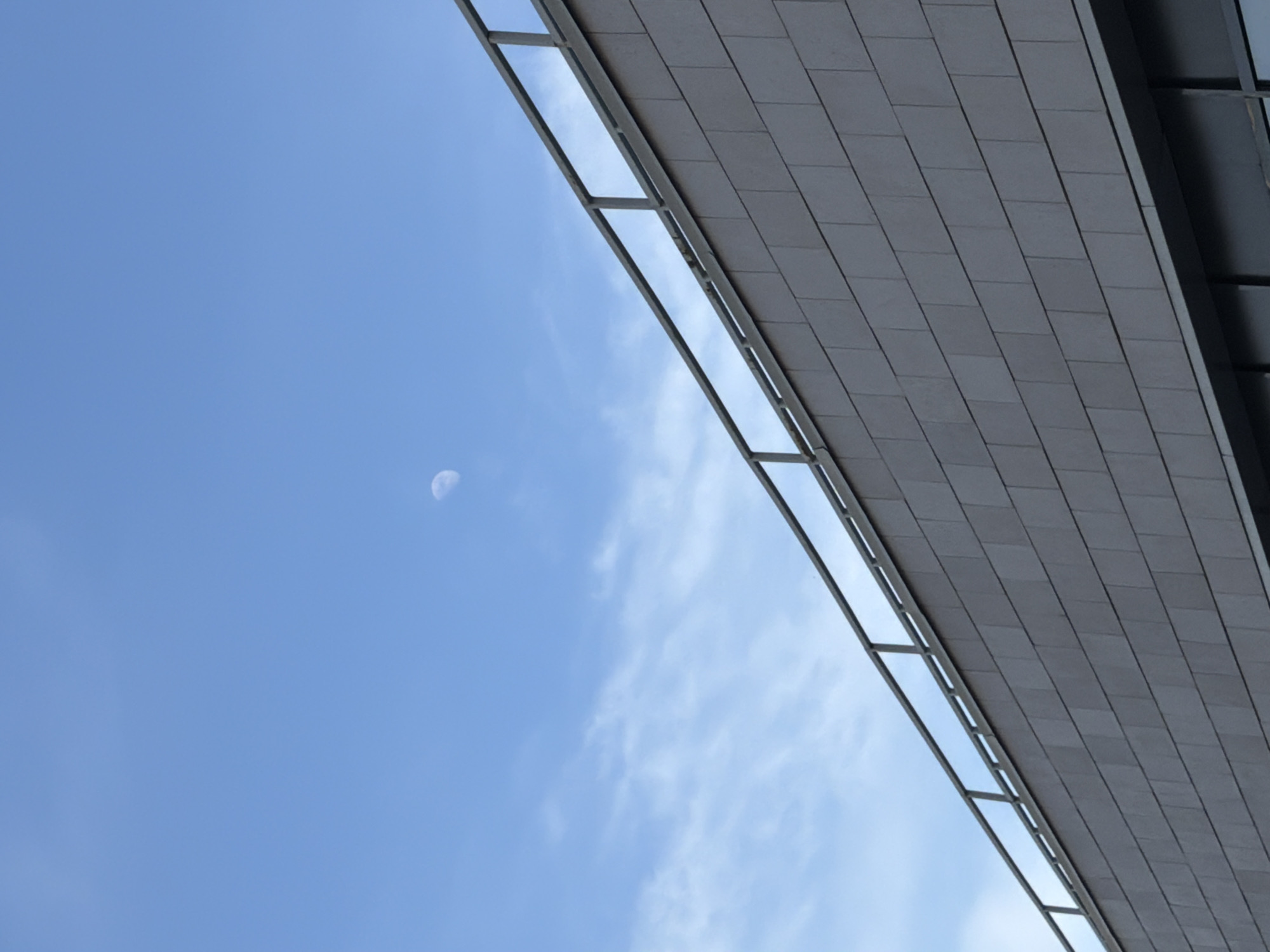The image shows a low-angle view of a building's exterior against a daytime sky in Krasnoyarsk, Russia. The building is clad in rectangular gray tiles. A metal framework runs along the edge of the building. The sky is a clear blue with thin clouds. The moon is visible in the upper-left quadrant.