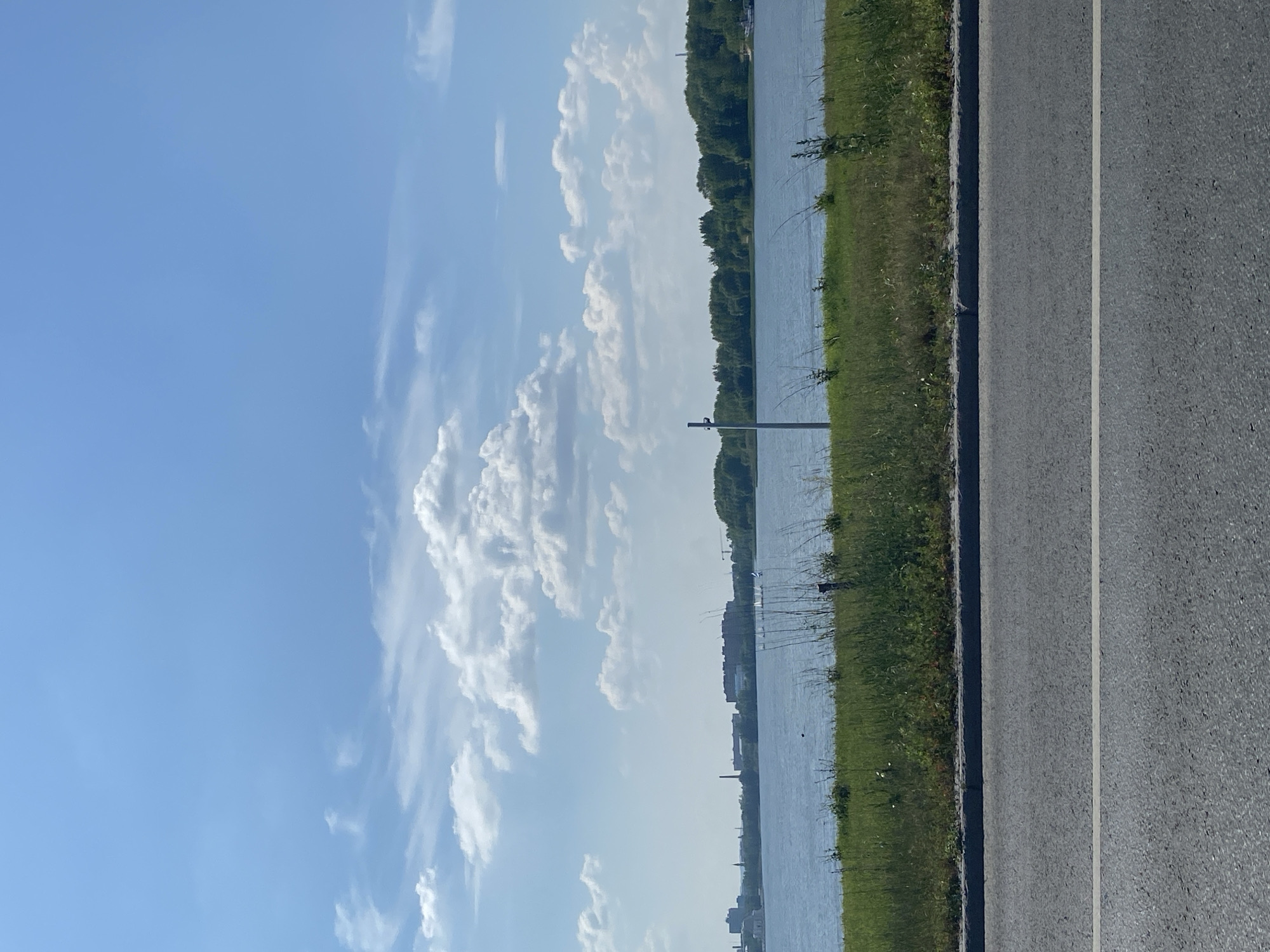 The image shows a body of water with a treeline on the far bank and a cityscape visible in the distance on the left. The sky is blue with scattered white clouds. In the foreground, there is a strip of grass followed by a paved road with a white line. The scene is likely located in the city of Kazan, Russia, possibly near the Kazanka River or one of the city's reservoirs, given the presence of urban structures and natural elements. There are no people or animals prominently featured in the image, and no specific actions are depicted. The environment appears to be an outdoor setting on a clear day.
