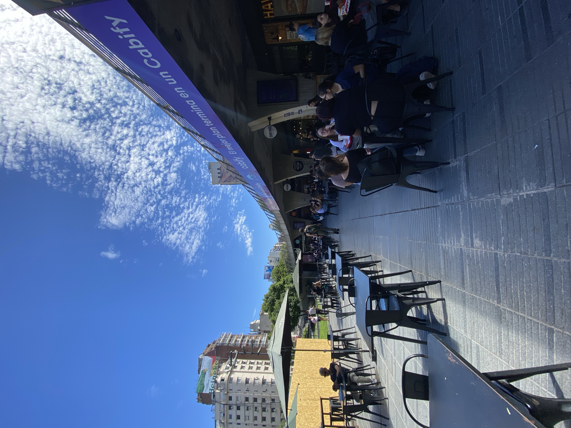 The image displays an outdoor dining area situated beneath a concrete viaduct in Buenos Aires, Argentina, consistent with the Paseo de la Infanta / Arcos del Rosedal area near the Rosedal de Palermo. The environment is illuminated by direct sunlight under a clear blue sky featuring scattered altocumulus clouds.
The foreground and midground are dominated by an array of empty and occupied black metal square tables with matching chairs, arranged on dark grey tiled pavement. Several dark green outdoor umbrellas are deployed, providing shade to various table settings. On the left, one male individual with dark sunglasses is seated alone at a table under an umbrella, engaging with a mobile phone.
The right side of the scene is defined by the underside of a substantial concrete viaduct, which supports a railway line, and houses multiple food establishments within its arches. Visible signage includes "TOSTADO," a partial McDonald's logo, and "HAVANNA." A long purple banner is affixed to the viaduct's underside, displaying "cabify El mejor plan termina en un Cabify" in white text.
Approximately 20-30 individuals are present. Groups of people are seated at various tables, engaged in conversation and consuming food or beverages. Other individuals are observed walking along the paved area; one person in camouflage trousers is seen walking towards the foreground in the midground.
The background on the left reveals multi-story urban buildings, one of which features a partially visible "Heineke" advertisement billboard on its upper facade. A segment of green trees and grass is visible between the buildings and the dining area.