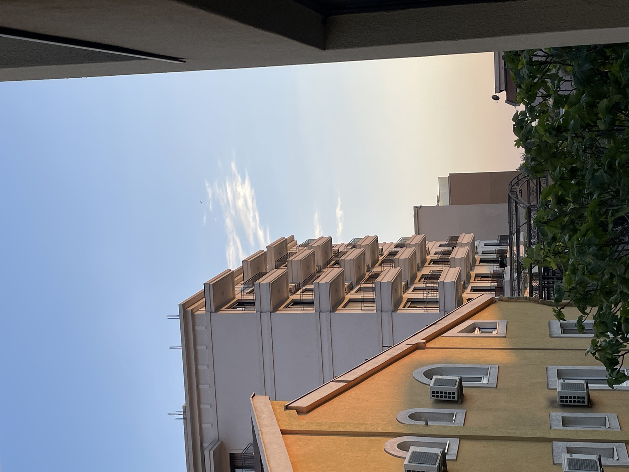 A low-angle shot depicts a building in Bar, Montenegro, viewed from within a building's frame. The sky is clear and blue. A tall white building with multiple balconies occupies the central left frame. A yellow building with rounded window frames and air conditioning units sits to the right. Green foliage fills the upper right.