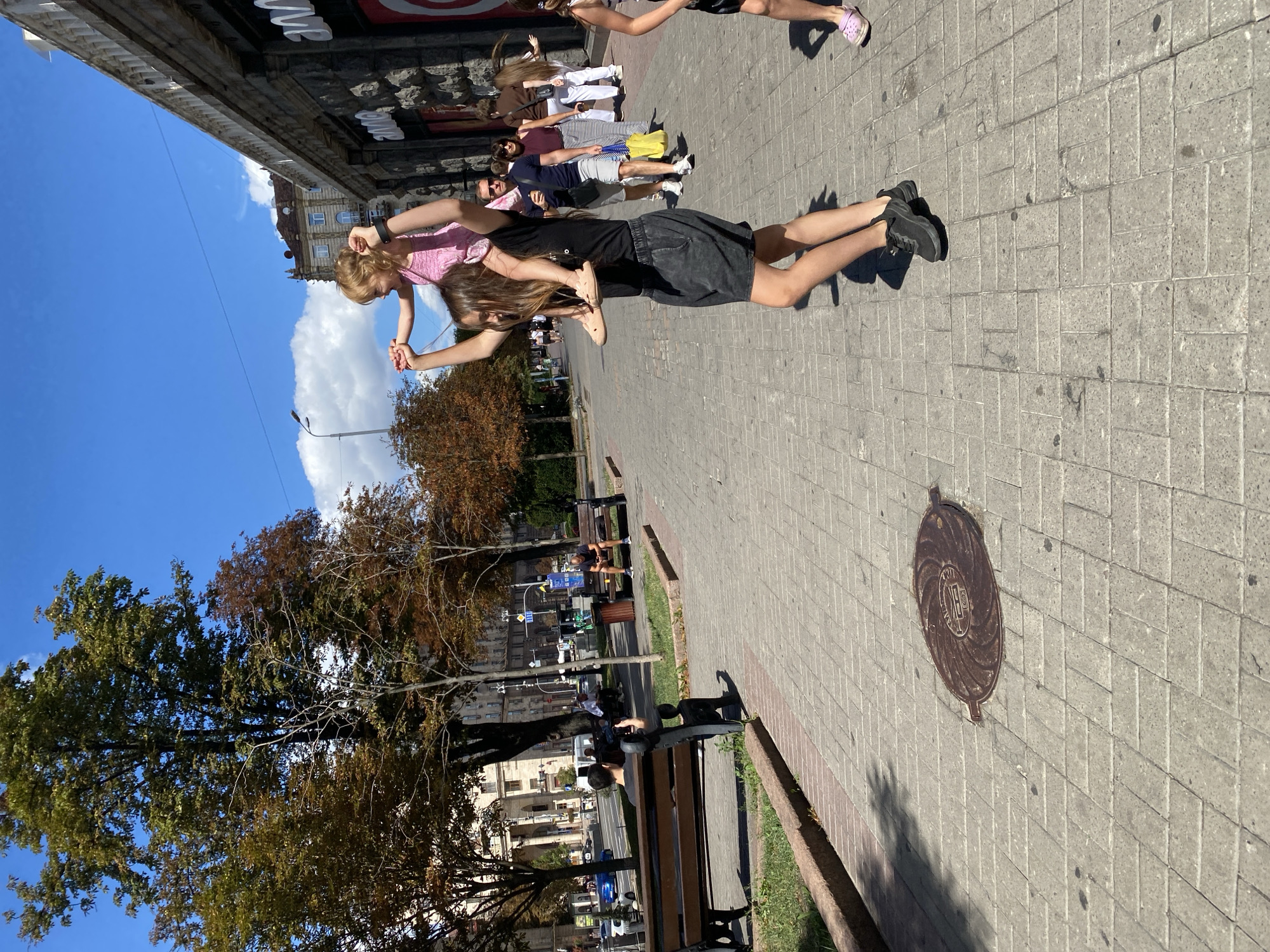 In an urban setting, a young woman with long brown hair, wearing a black t-shirt and dark grey shorts, carries a blonde child on her shoulders. The child is wearing a pink dress. They are walking on a paved sidewalk lined with trees and buildings. Several other people are visible in the background, some walking and others sitting on benches. The scene appears to be during the daytime, with sunlight casting shadows on the ground. There is a manhole cover on the sidewalk in the foreground.