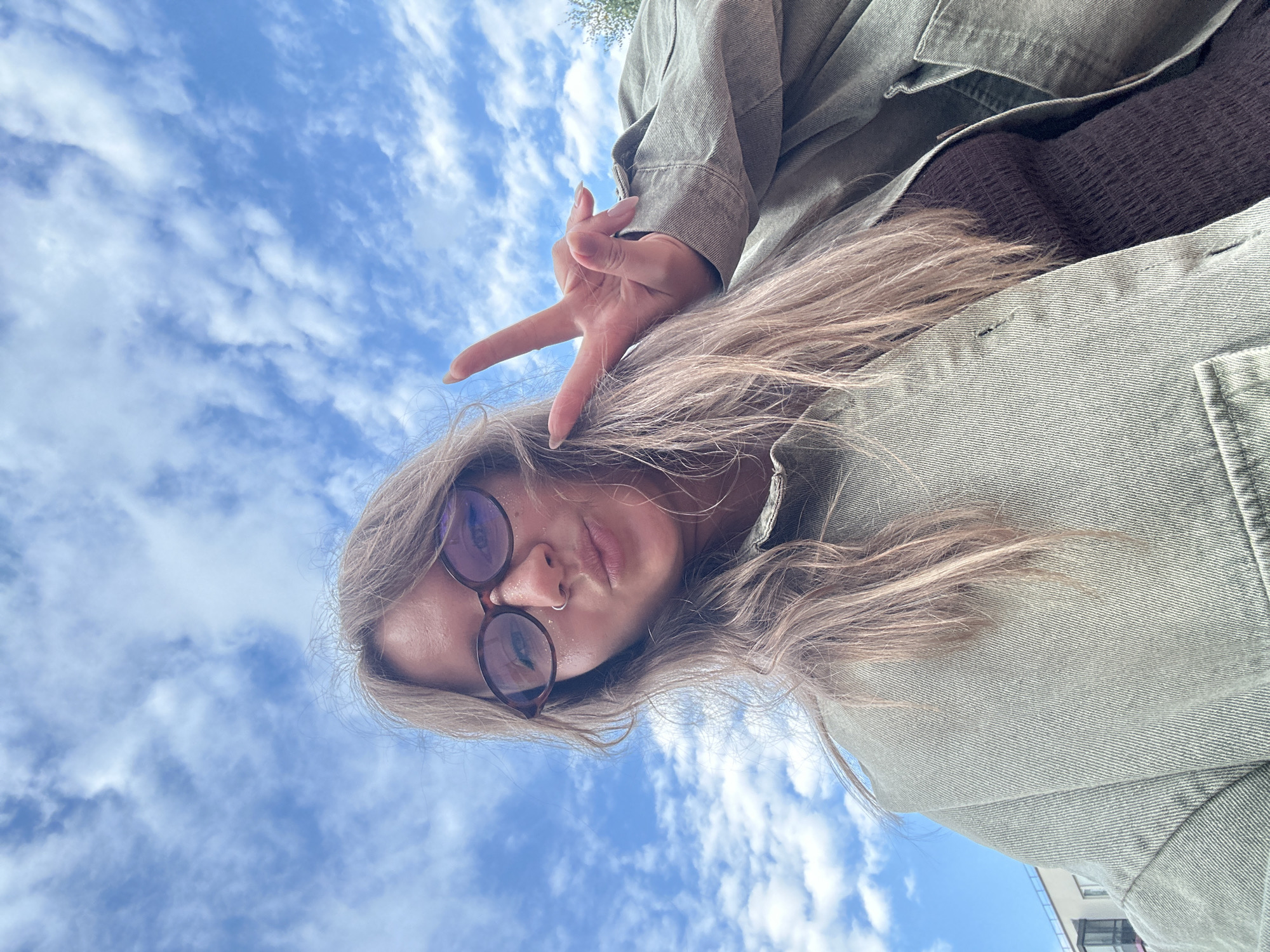 A young woman with blonde hair and glasses takes a selfie outdoors with a blue sky and white clouds in the background. She is wearing a light green jacket over a dark brown top and is holding up a peace sign with her right hand. The image appears to have been taken in Vsevolozhsk, Russia, as indicated by the presence of a modern apartment building in the background on the left side of the frame. The woman is positioned in the center of the frame, facing the camera with her mouth slightly puckered. Her hair is long and wavy, and she is wearing a nose ring. The lighting suggests it is daytime.