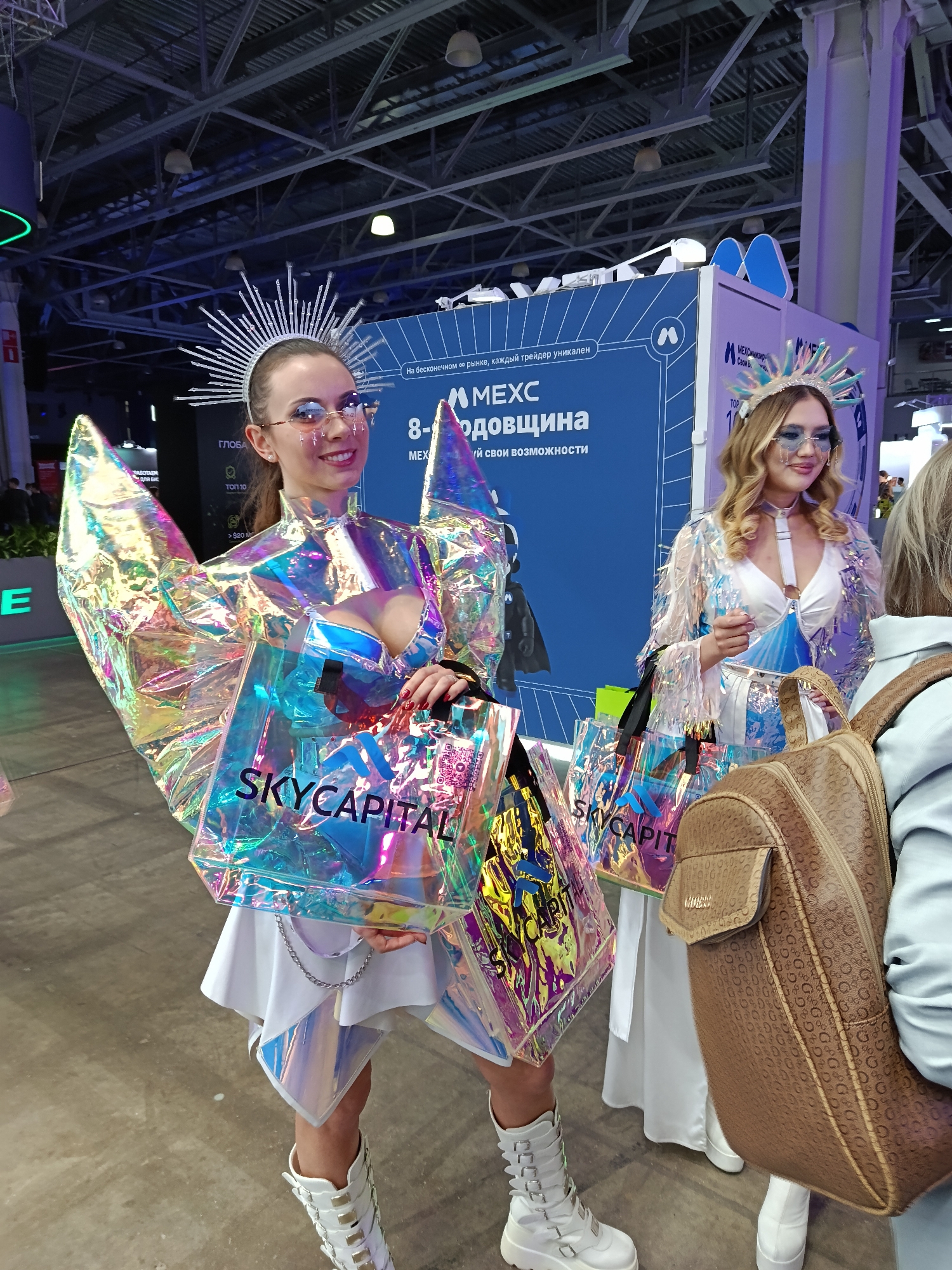 The image shows two women, likely models or promotional staff, at an event or convention in Moscow, Russia. The woman in the foreground is wearing a striking, iridescent costume with a starburst headpiece, white dress, and knee-high white boots. She is holding two clear shopping bags with "SKYCAPITAL" printed on them. The second woman is similarly dressed in an iridescent jacket, white dress, and a crown. A large blue backdrop with text in Russian, including "MEXC" and "8-одовщина" (8th Anniversary), is behind them. Part of a brown backpack is visible on the right side of the frame, held by an unseen person. The location appears to be a large convention center, with visible ceiling structures and convention booths in the background.