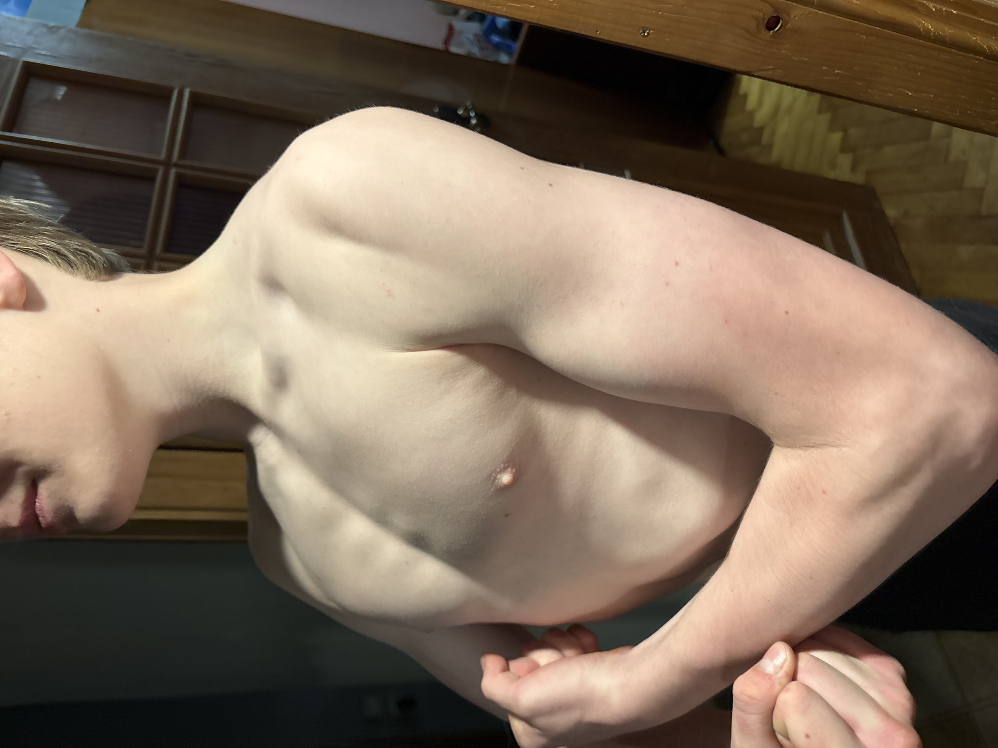 The image shows a shirtless young male, likely a teenager, posing with his arms flexed. The subject has fair skin and light-colored hair, partially visible at the top of the frame. He is wearing dark bottoms, likely shorts or pants. His chest and arm muscles are defined, suggesting physical fitness. The lighting in the image is dim, creating shadows that highlight the musculature.
The background consists of a wooden door with glass panels and a patterned wooden floor. The environment appears to be indoors, possibly a home. There are no visible elements that definitively place the scene in a specific city or country, let alone "Unknown, Russia." The image focuses solely on the subject and his immediate surroundings.