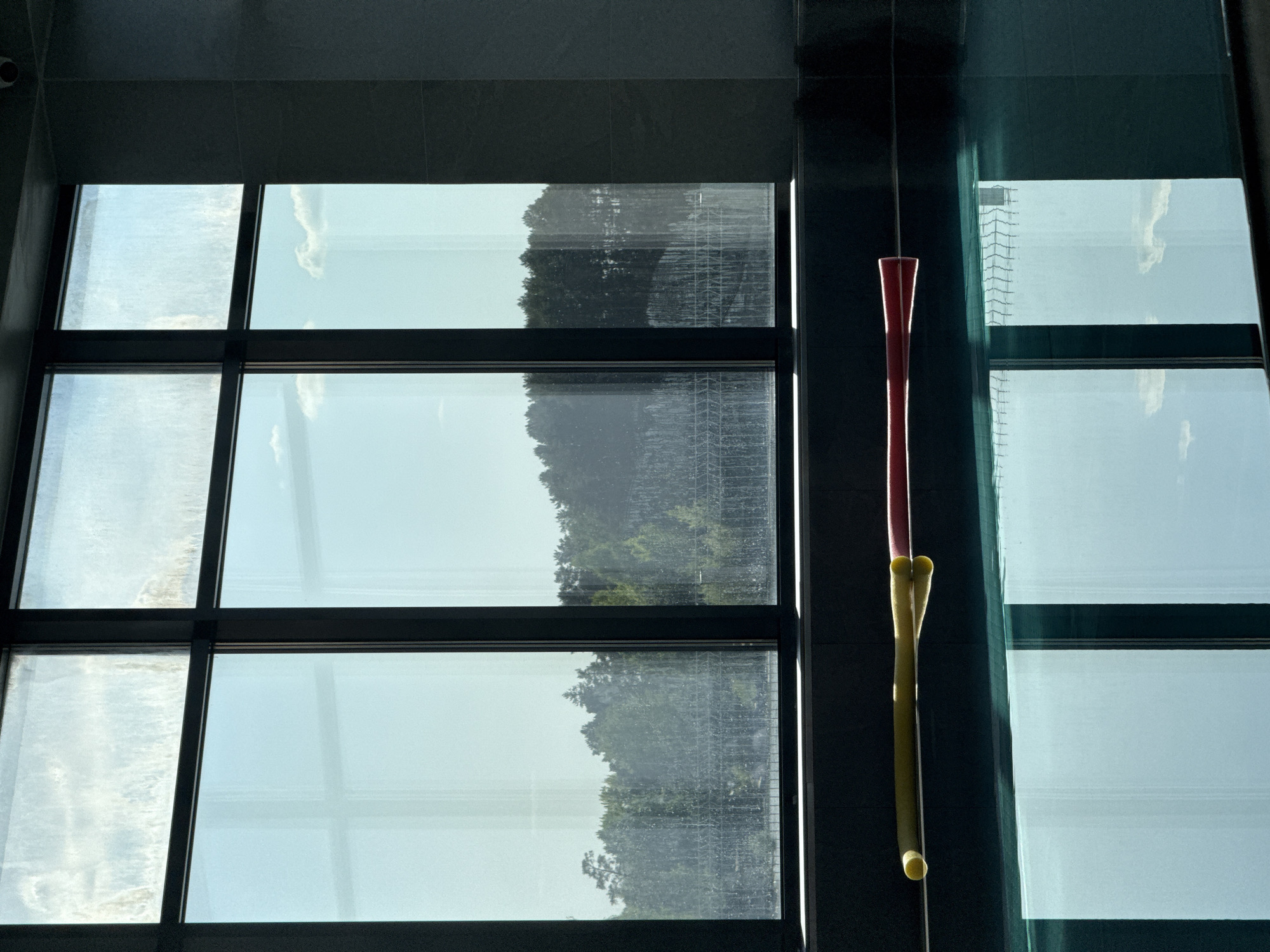 The image depicts a view through a large window with a black frame, divided into a grid of nine panes. Outside, trees and a light-colored sky are visible, slightly obscured by a film on the glass, possibly dirt or condensation. To the right of the window, an object hangs from the ceiling. It consists of a red, elongated shape at the top, connected to two yellow cylinders stacked on top of each other. The surrounding architecture includes dark walls. The location is Unknown.