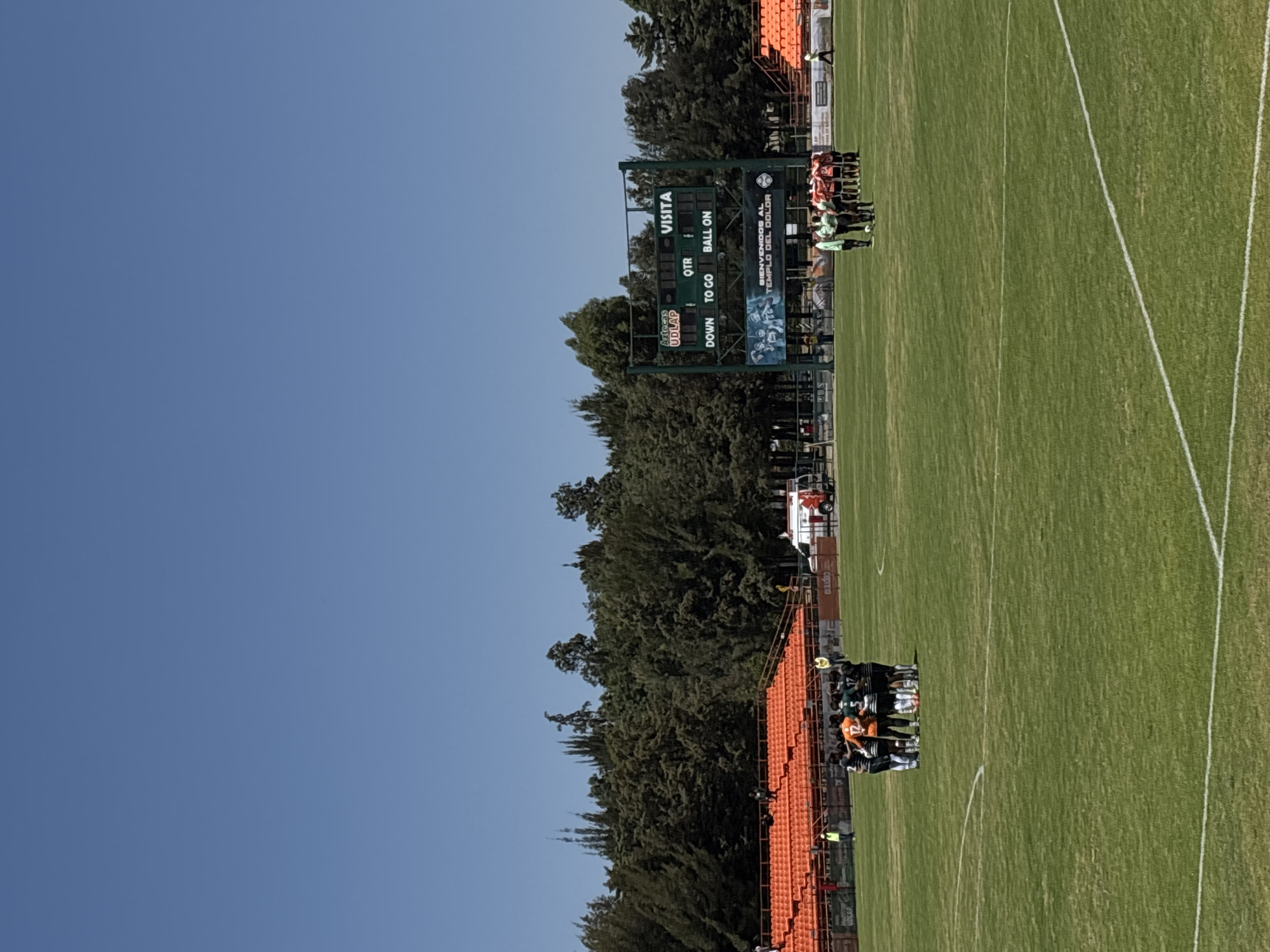 The image depicts an outdoor American football stadium, identified as the "Templo del Dolor" stadium of the Universidad de las Américas Puebla (UDLAP) in San Andrés Cholula, Mexico, based on text visible on the scoreboard and a banner. The scene is under a clear, bright blue sky. A green grass playing field with white line markings occupies the foreground and midground.
Two distinct groups of individuals, approximately 8-10 per group, are positioned in huddles on the field. The group on the left wears predominantly dark uniforms, some with black and white stripes, and one individual in an orange jersey with the number "12" visible. The group on the right wears mostly red or orange uniforms. One individual in a light green shirt stands just outside the right huddle, facing its members.
In the background, bright orange tiered spectator stands are visible on both sides of the field, appearing largely unoccupied. A white emergency vehicle (ambulance) with red markings is parked behind the right-side stands. A large green electronic scoreboard is centrally located behind the field. The scoreboard displays "Aztecas UDLAP" and "VISITA" at the top, along with fields for "QTR," "DOWN," "TO GO," and "BALL ON." A banner stretched below the main scoreboard reads "BIENVENIDOS AL TEMPLO DEL DOLOR." A dense line of green evergreen trees forms the outermost background behind the stadium structures. Several individuals in yellow vests are positioned near the stands.