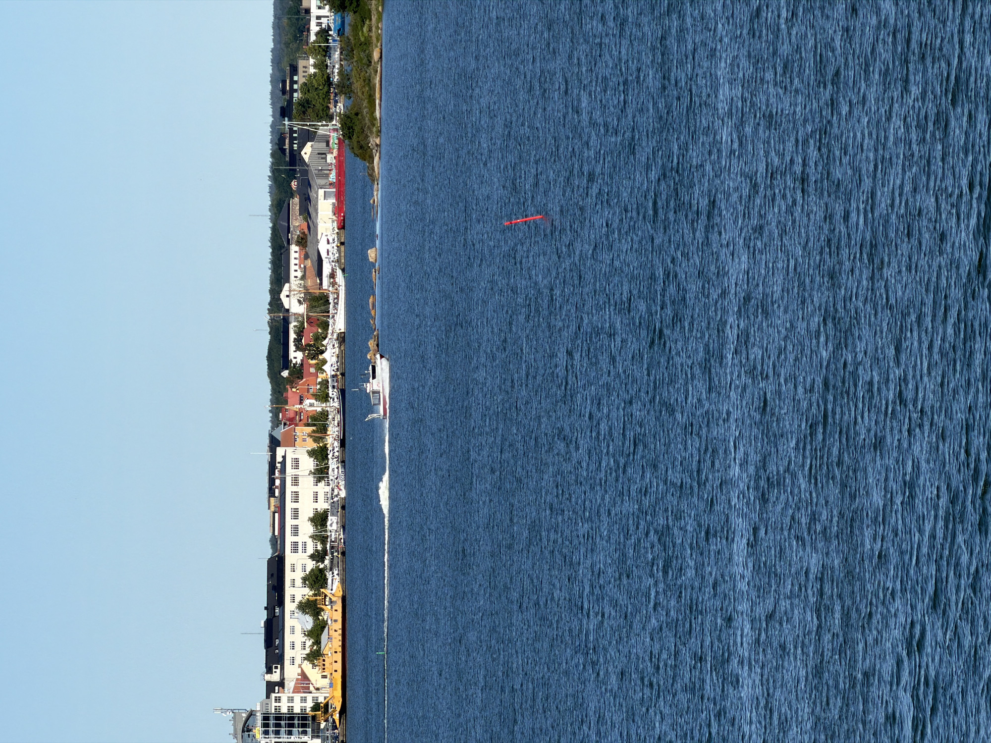 The image depicts a scene of the harbor in Karlskrona, Sweden, on a clear day with a bright blue sky. In the foreground, the deep blue water of the harbor is choppy with visible waves. A red buoy stands out in the middle of the water. A white and red boat is seen moving from left to right, leaving a white wake. In the midground, the shoreline is lined with buildings, including a tall modern building with glass panels on the left, followed by a row of older, multi-story buildings in various colors, predominantly white and yellow. Several boats are docked along the shoreline. Further in the background, more buildings and trees can be seen. The overall impression is of a busy maritime city on a sunny day.