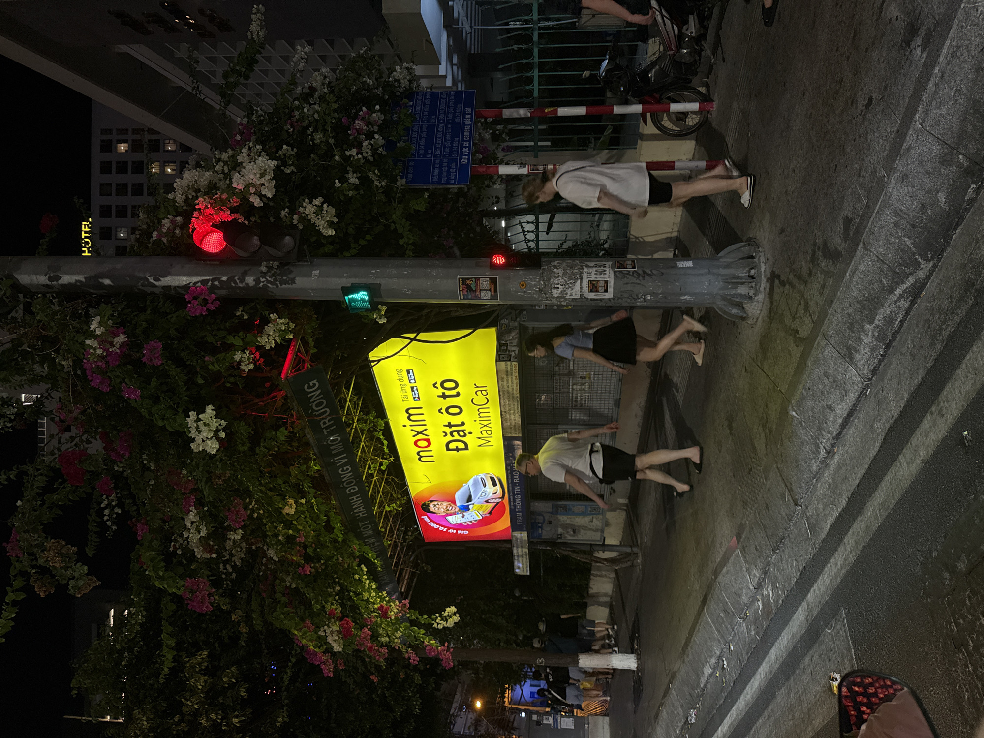 The image captures a street scene in Nha Trang Ward, Vietnam, at night. A prominent yellow advertisement for "maxim Đặt ô tô MaximCar" (Maxxim Book a Car MaxximCar) is affixed to a streetlight pole adorned with flowering vines. The pole also features a traffic light with a red signal and a green signal. A blue regulatory sign is visible above the traffic light.
Several pedestrians are present. One person is walking in shorts and a white shirt. Two more people are walking near the curb.
The surroundings feature buildings, including a hotel with the word "HOTEL" illuminated in yellow, indicating a business or lodging establishment. A red and white barrier is visible along the sidewalk, along with parked scooters. The street surface is dark and damp.