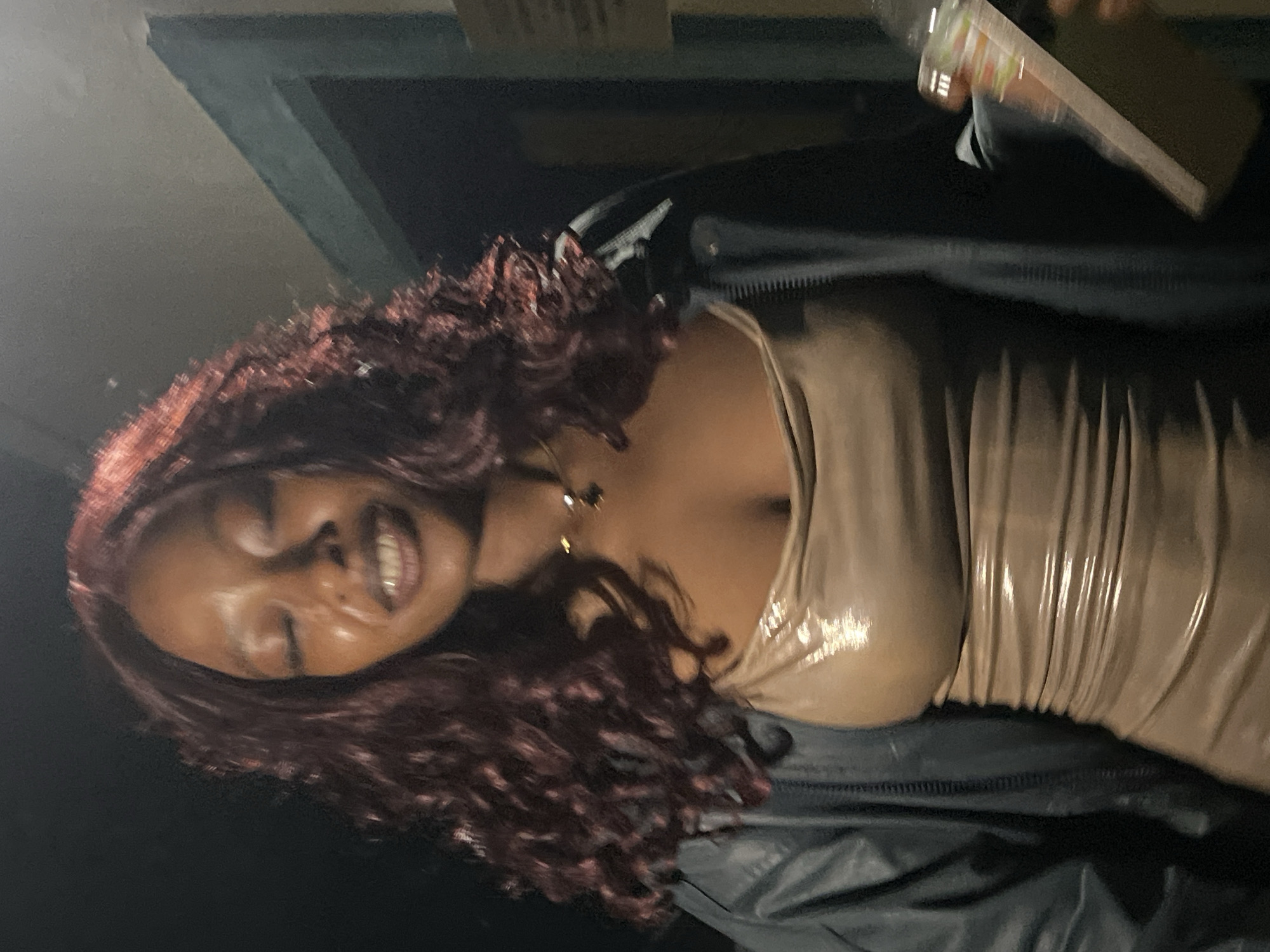 A medium shot captures a Black woman with long, dark, curly hair that appears to be a wig. She is wearing a tight, shiny, taupe-colored slip dress and a dark, possibly leather, jacket. Her eyes are closed, and her mouth is open in a smile, revealing her teeth. She is wearing a delicate necklace with a dark pendant. Her right hand, partially visible, holds a clear plastic cup with a straw. The background is dark and indistinct, with a hint of a doorway or frame. The image appears to be taken indoors or at night, with the lighting focused on the woman. There is no identifiable information regarding the specific location within the city of Durimi, Nigeria.