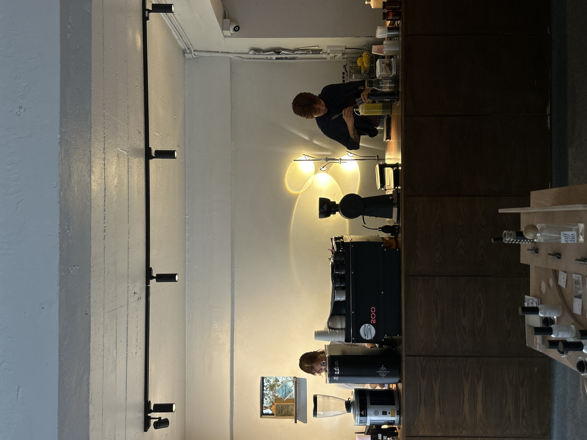 The image depicts the interior of a coffee shop, likely in Chiang Mai City Municipality, Thailand, characterized by its modern, minimalist design. Two baristas are working behind a wooden counter. On the left, a barista with reddish-brown hair is partially obscured by a coffee grinder and an espresso machine. To the right, another barista, also with reddish-brown hair, is focused on preparing a drink, stirring a yellow liquid in a container. The counter displays various coffee-making equipment, cups, bottles, and what appears to be a fruit basket containing lemons. Above the counter, track lighting illuminates the space, and a distinctive circular lamp creates a warm glow on the wall behind the second barista. A small, framed picture hangs on the wall above the espresso machine. In the foreground, a wooden table is set with several bottles, likely nail polish, and other small items, suggesting it might be a display or a waiting area. The overall ambiance is calm and focused, with an emphasis on clean lines and functional aesthetics.