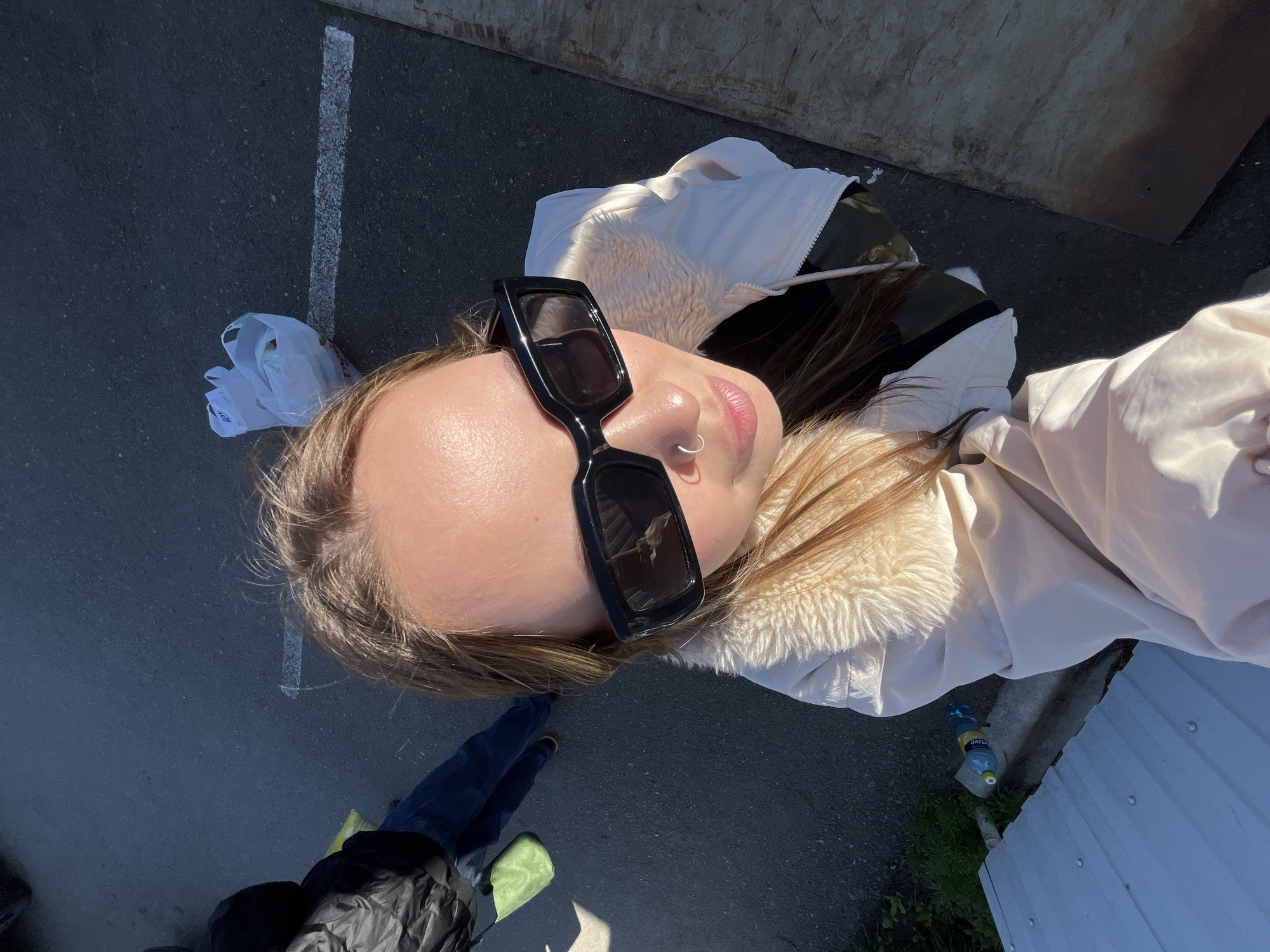 A selfie is taken from a low angle of a young woman wearing black sunglasses, a nose ring, and a cream-colored jacket with a fur collar. She is standing outdoors, on a paved surface with a white line and a plastic bag visible on the ground. Another person, wearing dark pants and a dark jacket with a light-colored bag, is partially visible at the bottom left of the frame. In the background, there is a metal sheet and what appears to be a bottle of water on the ground near a wall. The location might be near Novopokrovskaya, Russia.