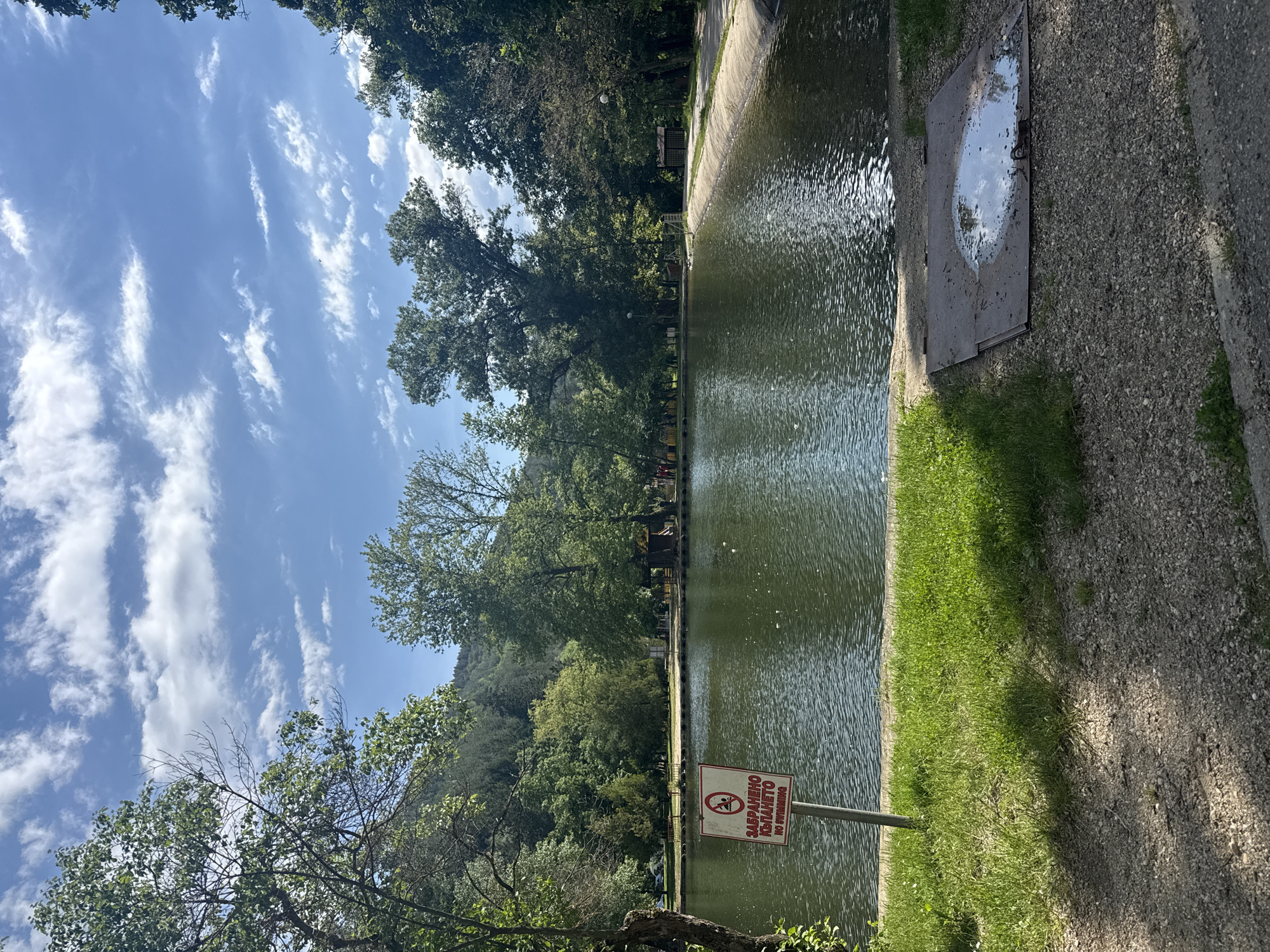 The image shows a long, narrow body of water in a park-like setting. On the right side is a concrete edge, then a strip of grass, then a gravel path. A metal access cover is partially submerged in the water near the edge. A sign, written in Bulgarian and English, indicates that swimming is prohibited. Trees line the far side of the water. The sky is blue with streaks of clouds. The location is most likely a channel of the Blagoevgradska Bistritsa River in Blagoevgrad, Bulgaria.