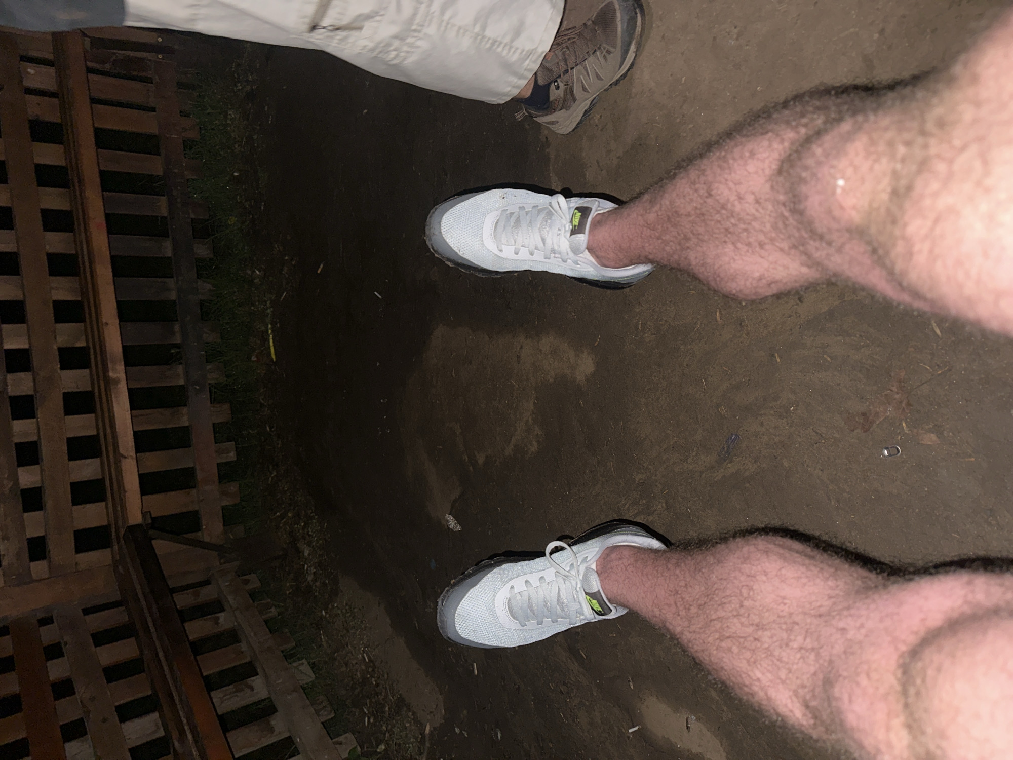 This overhead shot depicts the lower bodies of two individuals standing on dark, earthy ground, likely outdoors at night in Blagoevgrad, Bulgaria, given the low light and artificial illumination.
In the center and foreground, a person's two hairy legs are prominent, wearing light grey and white sneakers with green detailing, prominently featuring the "NIKE" logo on the side. The ground beneath their feet is compacted earth or dirt, appearing damp or dark. A small, shiny, U-shaped metal object lies on the ground to the right of their left foot, and scattered small debris is visible.
In the upper part of the image, the lower leg of a second person is visible, clad in light-colored trousers (possibly khakis) and a dark outdoor shoe, which appears to be a hiking boot.
To the left, a wooden slatted structure, resembling stacked pallets or a wooden barrier, is partially visible, with sparse green grass growing between it and the dirt surface. The dim lighting creates distinct shadows from the legs and shoes, indicating nighttime conditions. No specific activity beyond standing or walking is discernible.
