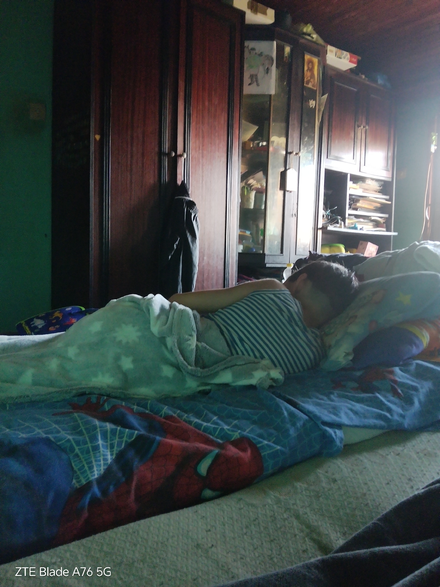 In a dimly lit bedroom, a person lies asleep on a bed covered with a Spider-Man themed comforter and a white blanket with a star pattern. The person is wearing a striped t-shirt. Behind the bed, a large, dark wooden wardrobe with several doors stands against a green wall. The wardrobe has some items displayed on shelves within it, including pictures and books. The overall impression is of a personal space, likely a home in Stara Zagora, Bulgaria.