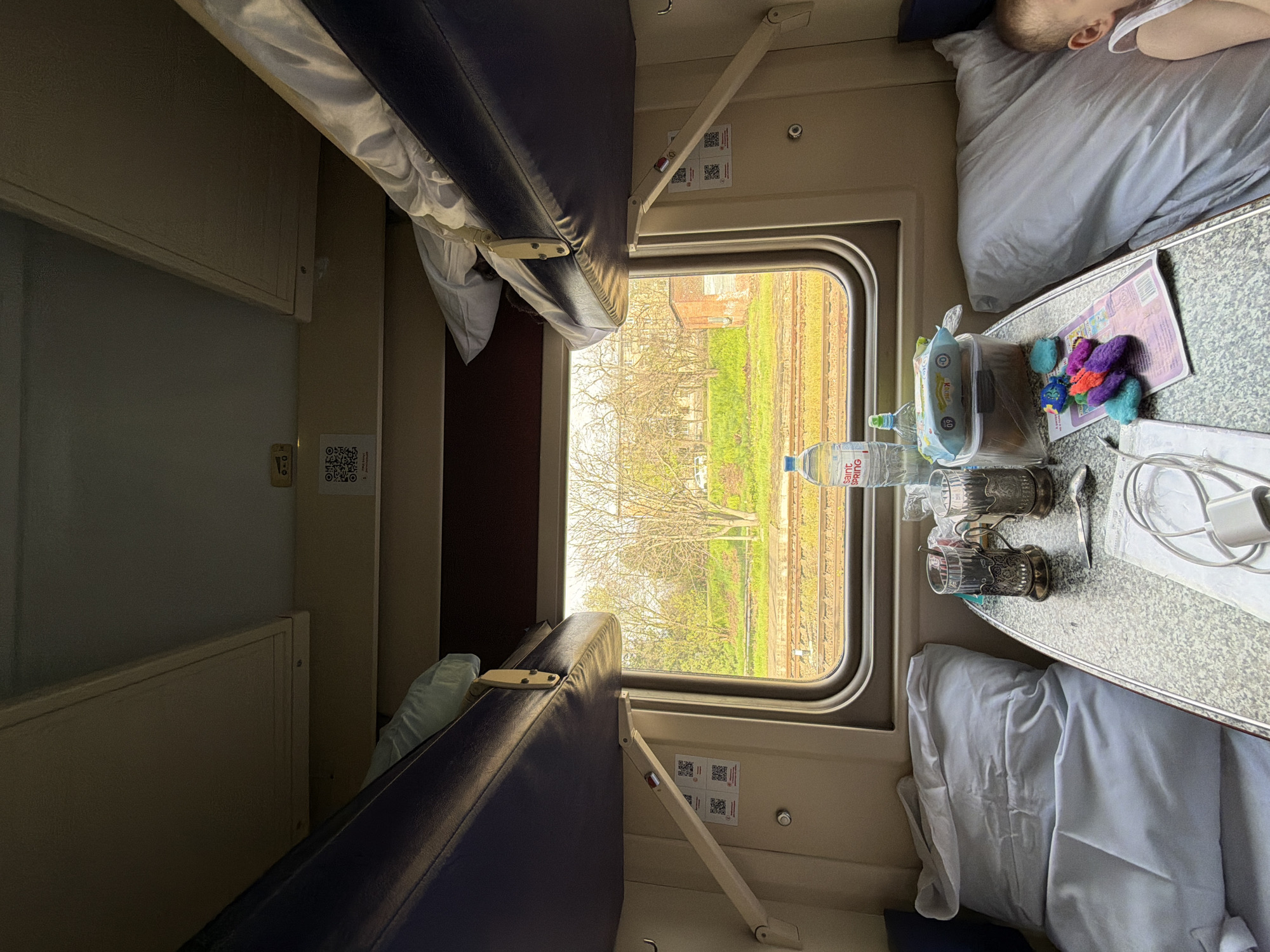 The image depicts the interior of a train compartment. There are two beds on either side of the compartment, one raised and one lowered. A person is lying on the lower bed on the right side, with only the side of their face visible. Between the bed and the window, there is a small table holding a water bottle, a container of baby wipes, colorful toys, a book, two decorative glasses with spoons, and charging cables. Through the window, trees and vegetation can be seen, as well as railroad tracks. The compartment's walls are beige.