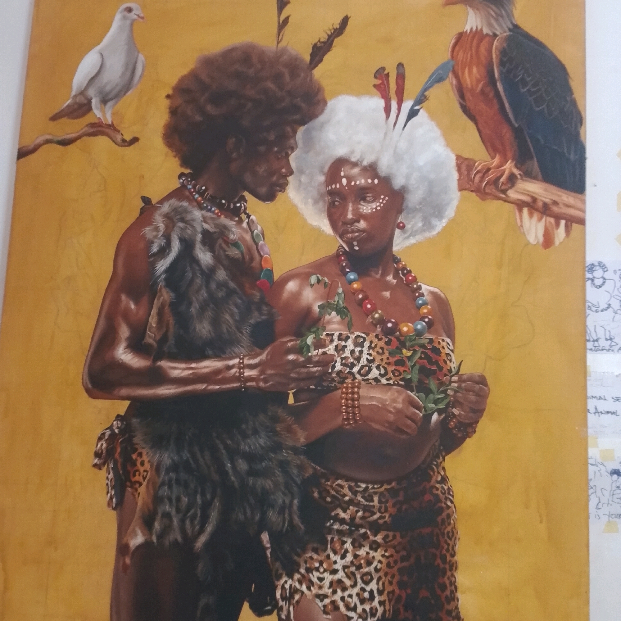 This is an oil painting depicting an indigenous couple in traditional attire against a yellow, textured background. The male figure on the left has dark skin, a large afro, and wears a necklace of colorful beads and a fur garment. He is embracing the female figure. The female figure on the right has dark skin, a white afro, and white markings on her face. She wears a similar colorful beaded necklace and a leopard-print garment. She holds a sprig of green leaves. To the left of the couple, a white dove perches on a branch. To the right, an eagle perches on another branch. The painting is a detailed representation of the subjects and their adornments. There is no identifiable location in the city of Ado, Nigeria within the image itself.
