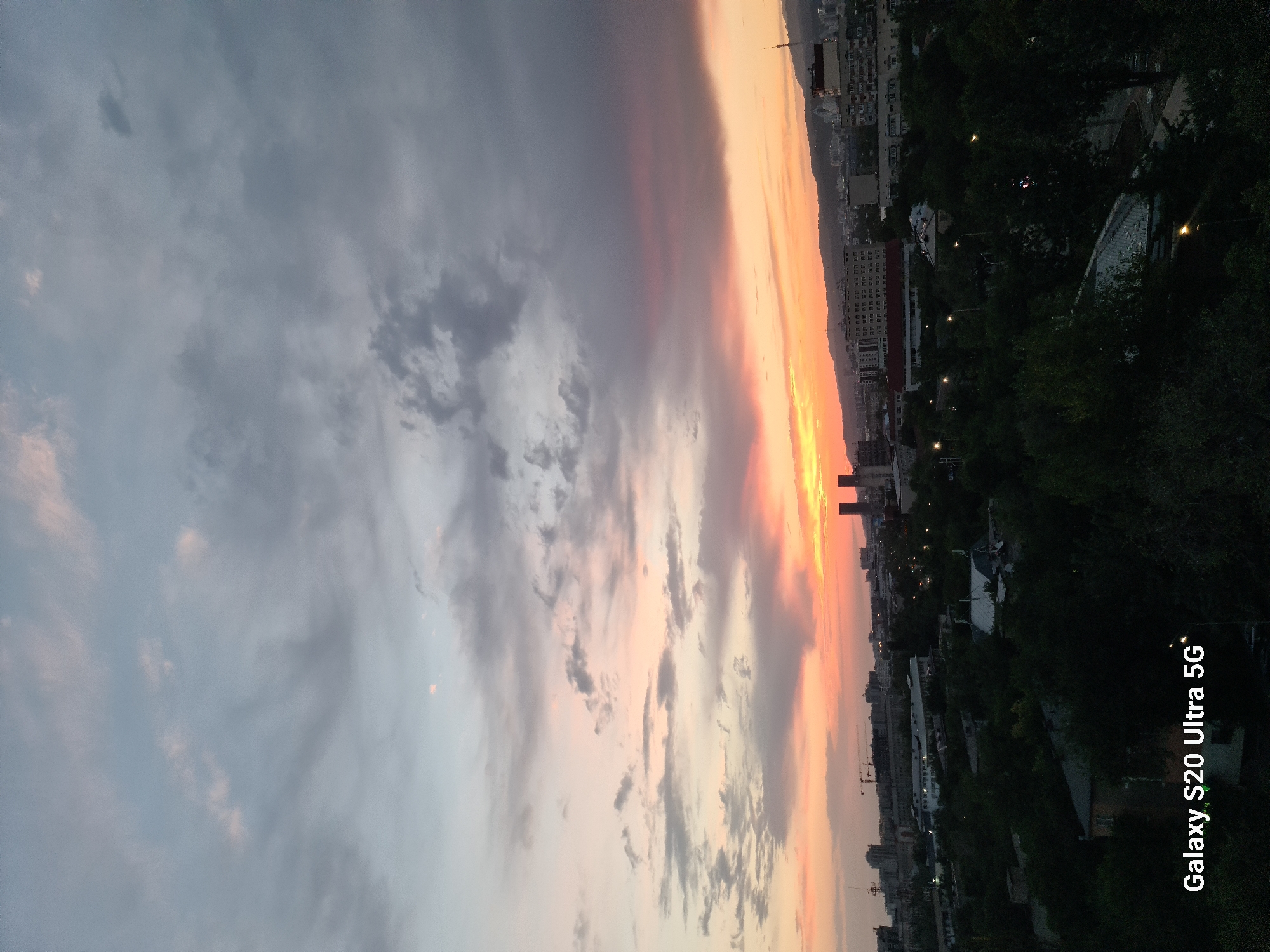 The image shows a city skyline at dusk with a vibrant sunset. The sky is filled with dramatic clouds tinged with orange, pink, and purple hues, indicating the sun is setting behind the mountains. In the foreground, there is a dense canopy of dark green trees, obscuring much of the ground level. Above the trees, a cityscape is visible with various buildings, including some taller structures and what appear to be construction cranes. In the distance, mountains form a backdrop to the city. The bottom left corner of the image contains a watermark reading "Galaxy S20 Ultra 5G". The scene is consistent with a view from an elevated position looking west over the city of Almaty, Kazakhstan, given the presence of mountains to the west and the typical urban architecture. There are no people or animals visible in the image.