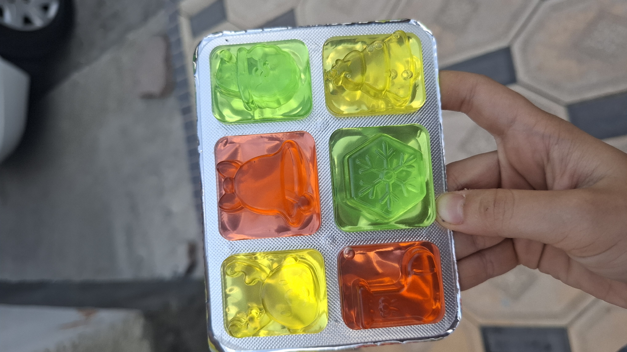 In Shymkent, Kazakhstan, a hand holds a blister pack of six Christmas-themed jelly candies. The candies are in two rows of three. The top row contains a yellow reindeer, an orange bell, and a green Santa Claus. The bottom row contains an orange stocking, a green snowflake, and a yellow Christmas tree. The blister pack is silver and has a textured surface. The background is blurred but appears to be an outdoor setting with a paved surface.