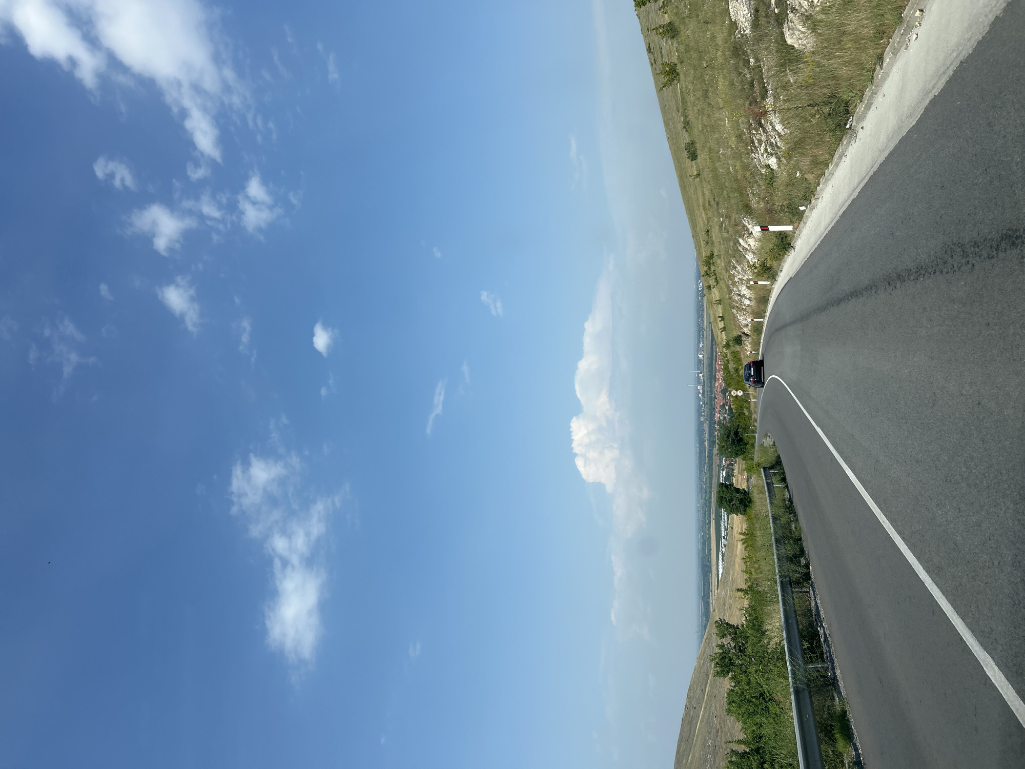 The image displays a clear daytime scene of a paved road winding through a hilly, semi-arid landscape near Isperihovo, Bulgaria. The road, dark asphalt with a solid white line on its left edge, curves right in the foreground and continues into the distance. A single dark-colored sedan is visible on the right side of the road, traveling away from the viewer. To the left of the road, a silver metal guardrail runs alongside a vegetated embankment of green bushes and dry, yellowish-brown grass leading up a hill. To the right, white and red reflective roadside markers delineate the edge of the road, followed by a concrete curb and then a rocky, grassy slope covered in dry vegetation and sparse small trees. In the middle ground, through the valley framed by the hills, a settlement identified as Isperihovo is visible, characterized by numerous buildings with red roofs. Beyond the settlement, industrial structures, possibly chimneys, and flat agricultural fields are discernible under a hazy sky. The sky above is a bright blue with scattered white cumulus clouds, particularly clustered near the horizon. The scene is brightly illuminated by natural sunlight, casting visible shadows.