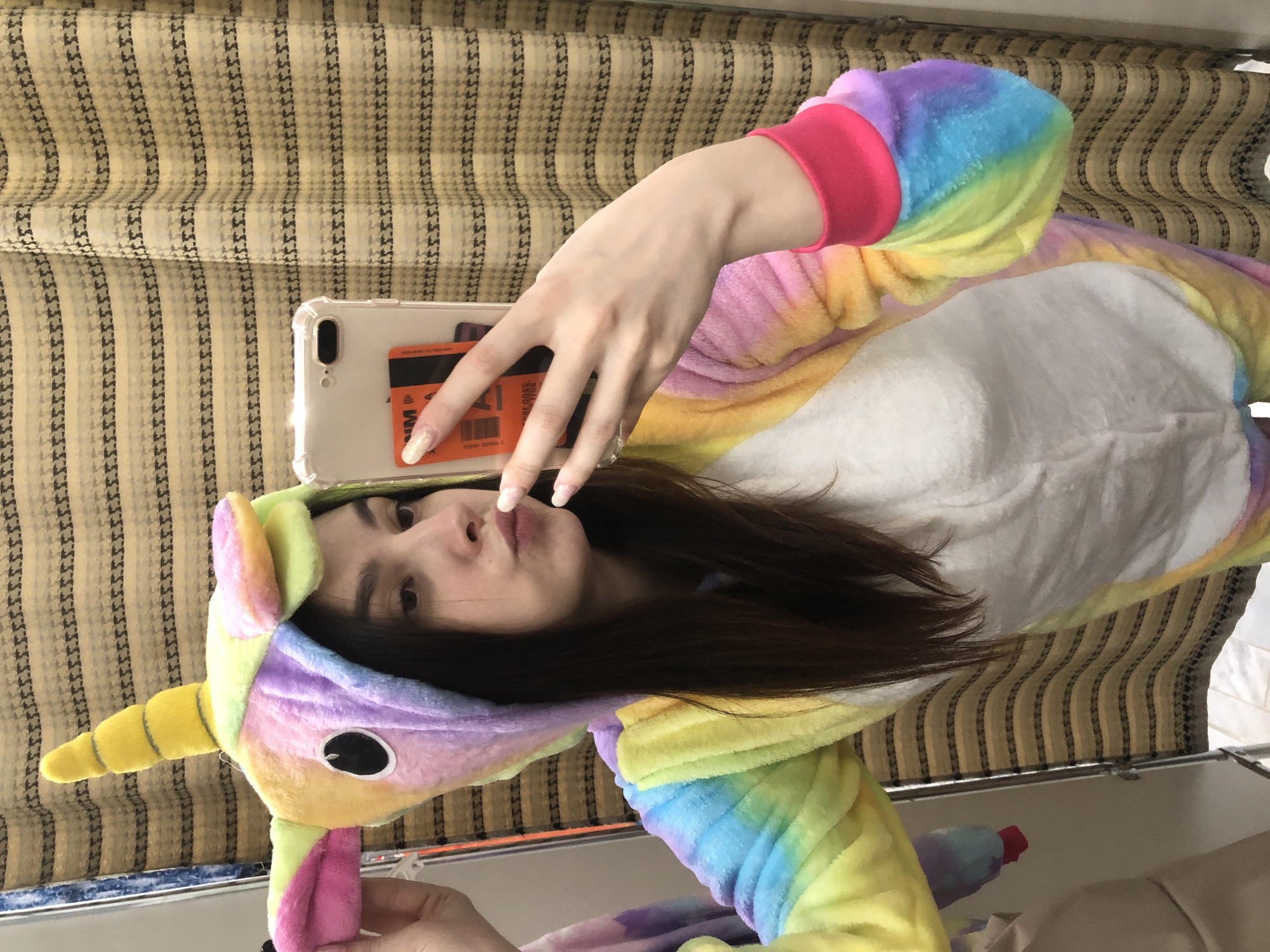 A person in a rainbow unicorn onesie is taking a selfie with a smartphone. The phone case appears clear with an orange card inserted. The person has long, dark hair and is indoors. The background includes a curtain with a striped pattern. The location of Chaikovsky, Russia cannot be determined from the image.