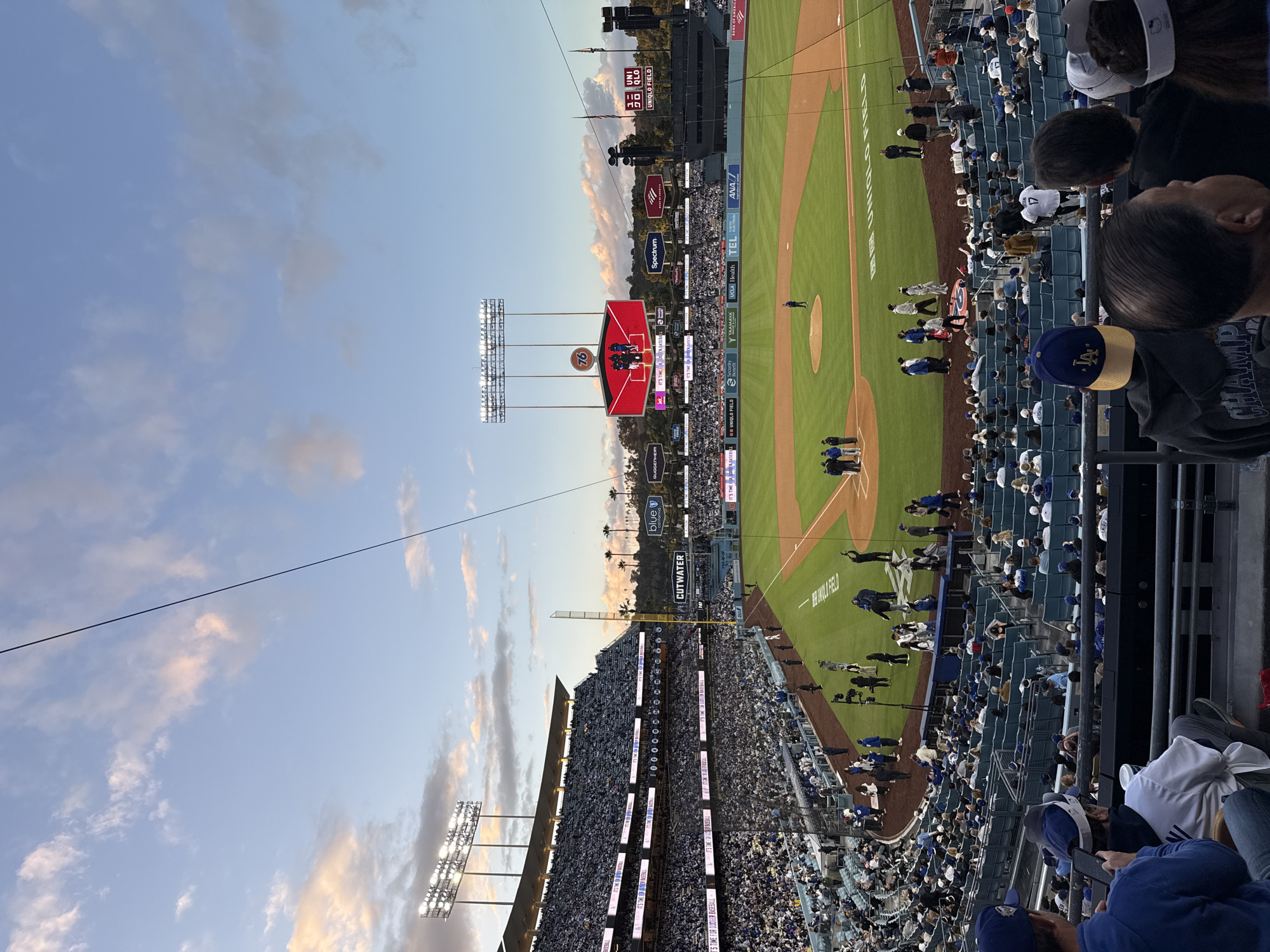 This image shows a baseball game in progress at Dodger Stadium, Los Angeles, California. The stadium is filled with spectators, many wearing blue Dodgers gear. The field is green, and players are visible on the field. The Jumbotron displays a baseball graphic, and advertisements for sponsors like Uniqlo, Spectrum, and UCLA Health are visible. The sky above the stadium is partly cloudy at sunset.